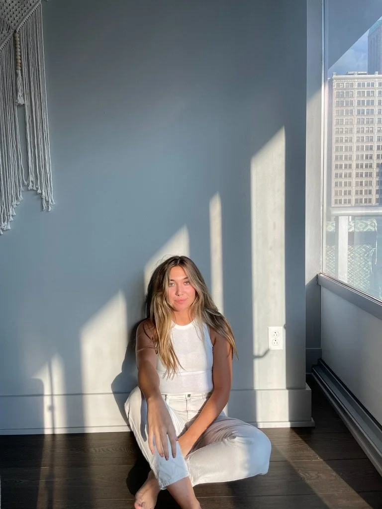 A woman sitting on dark wood floor next to a large window with sunlight casting shadows on the wall behind her.