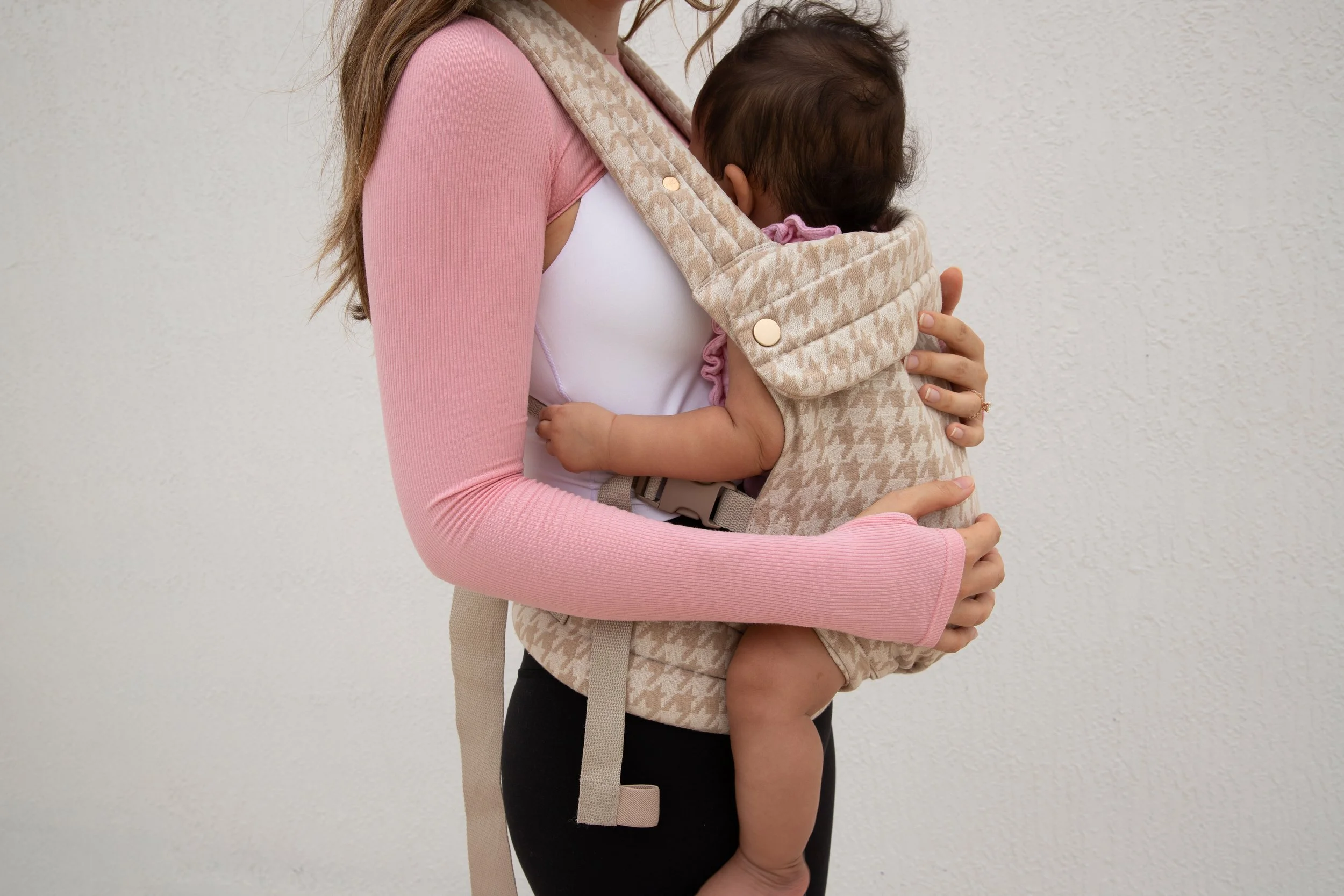 A woman wearing a pink long-sleeve shirt carries a baby in a beige houndstooth-patterned baby carrier against a plain white wall.