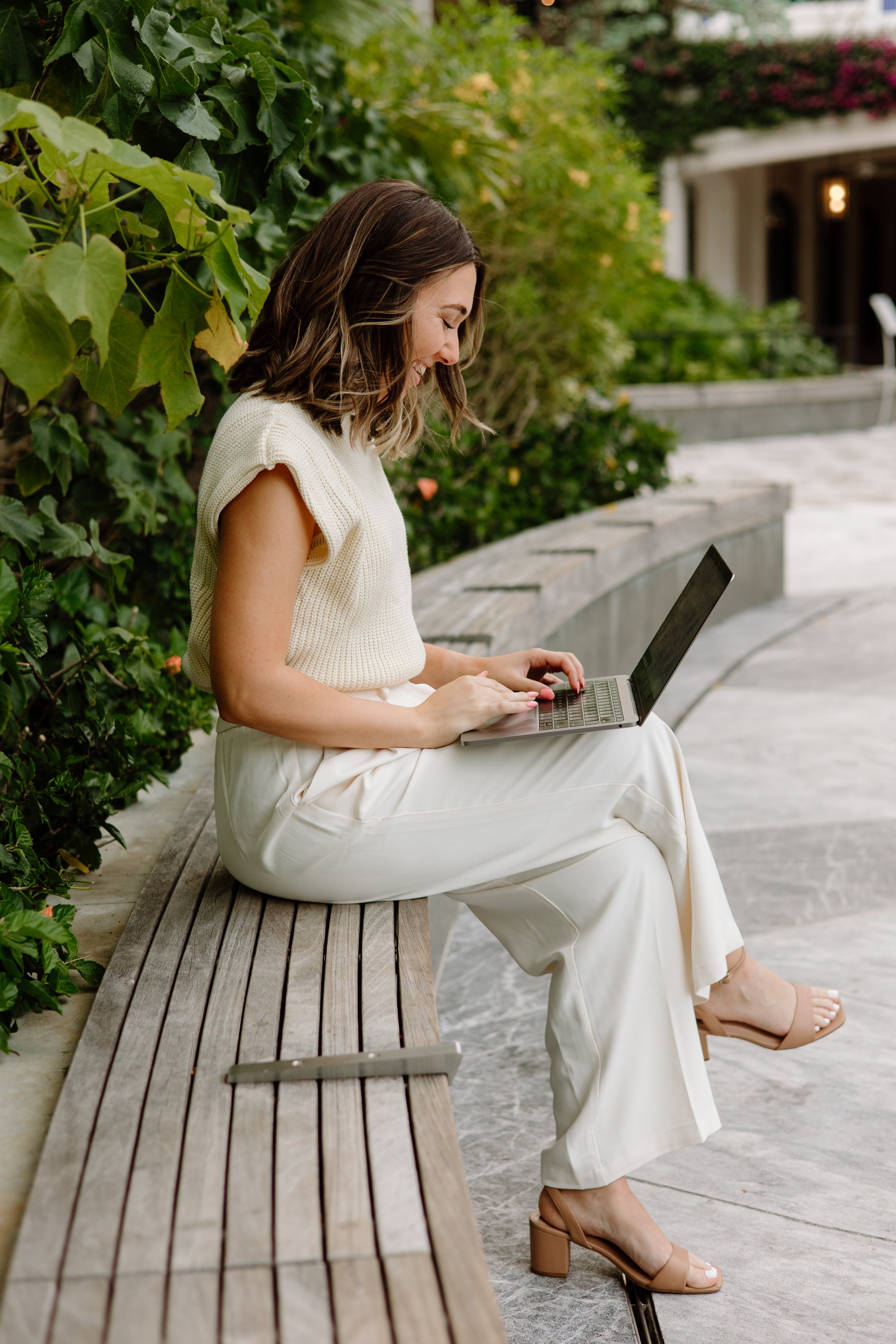 Woman sitting on a wooden bench working on a laptop outdoors, surrounded by green plants and flowers, during daytime.