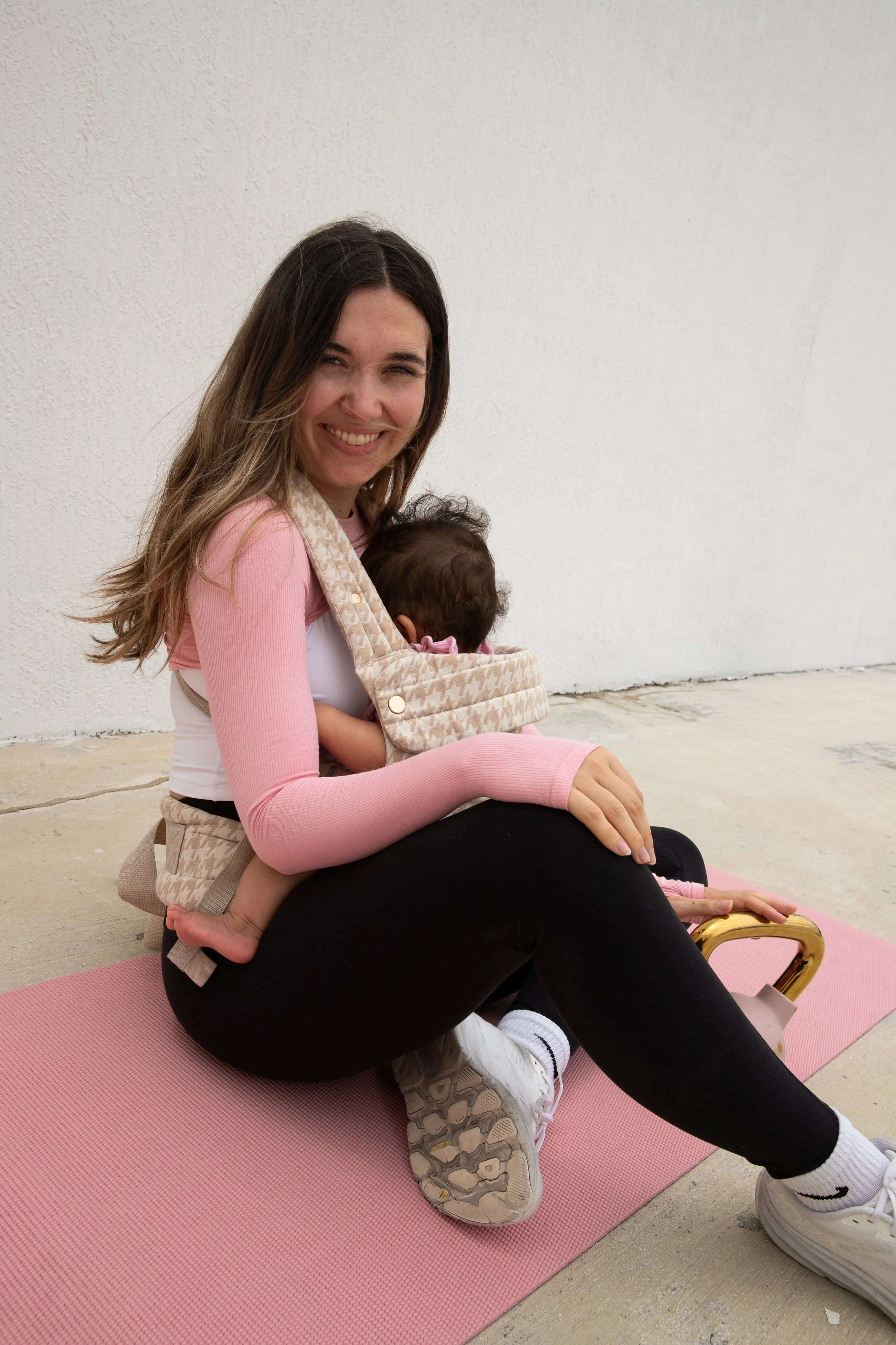 Young woman sitting on a pink yoga mat outside, holding a small child in a baby carrier, practicing yoga or stretching.