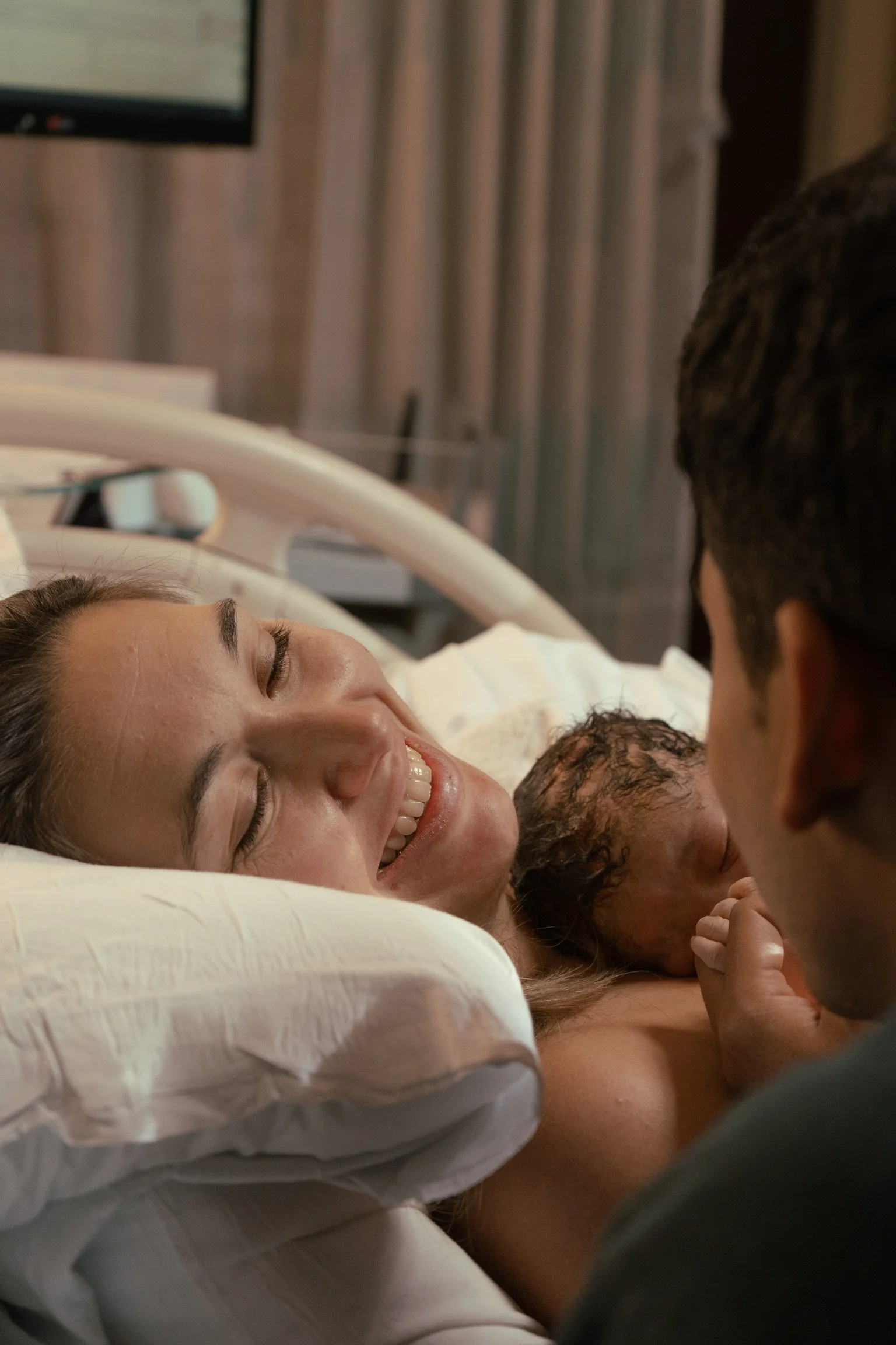 A woman lying in a hospital bed smiling while a man and a child, presumably her partner and child, are close to her. The woman appears to be in labor or just gave birth, and the man and child are touching her face and showing affection.