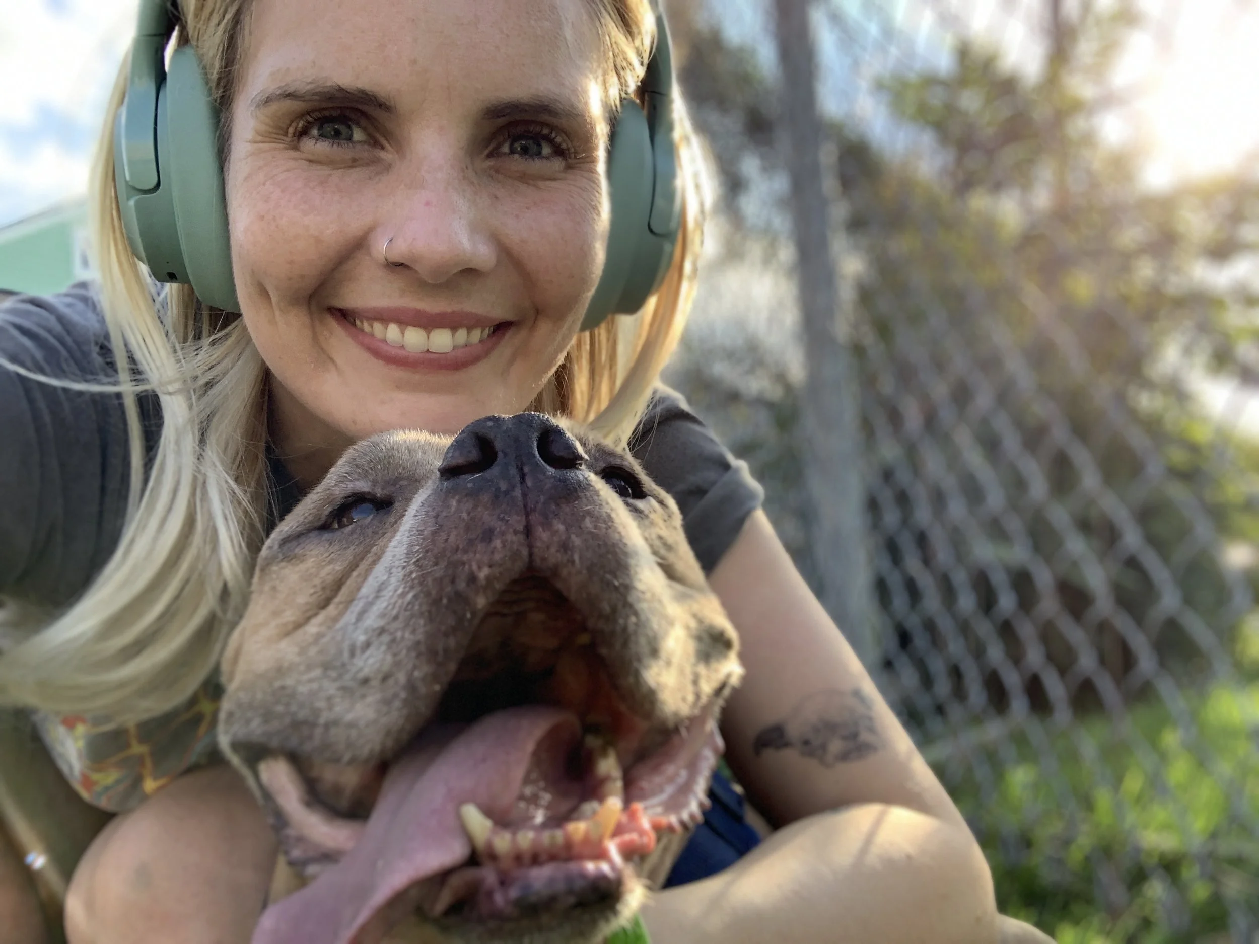 A woman with blonde hair, a nose ring, and a tattoo on her arm, smiling while holding a large, gray dog with a wide, open mouth, outdoors with trees in the background.