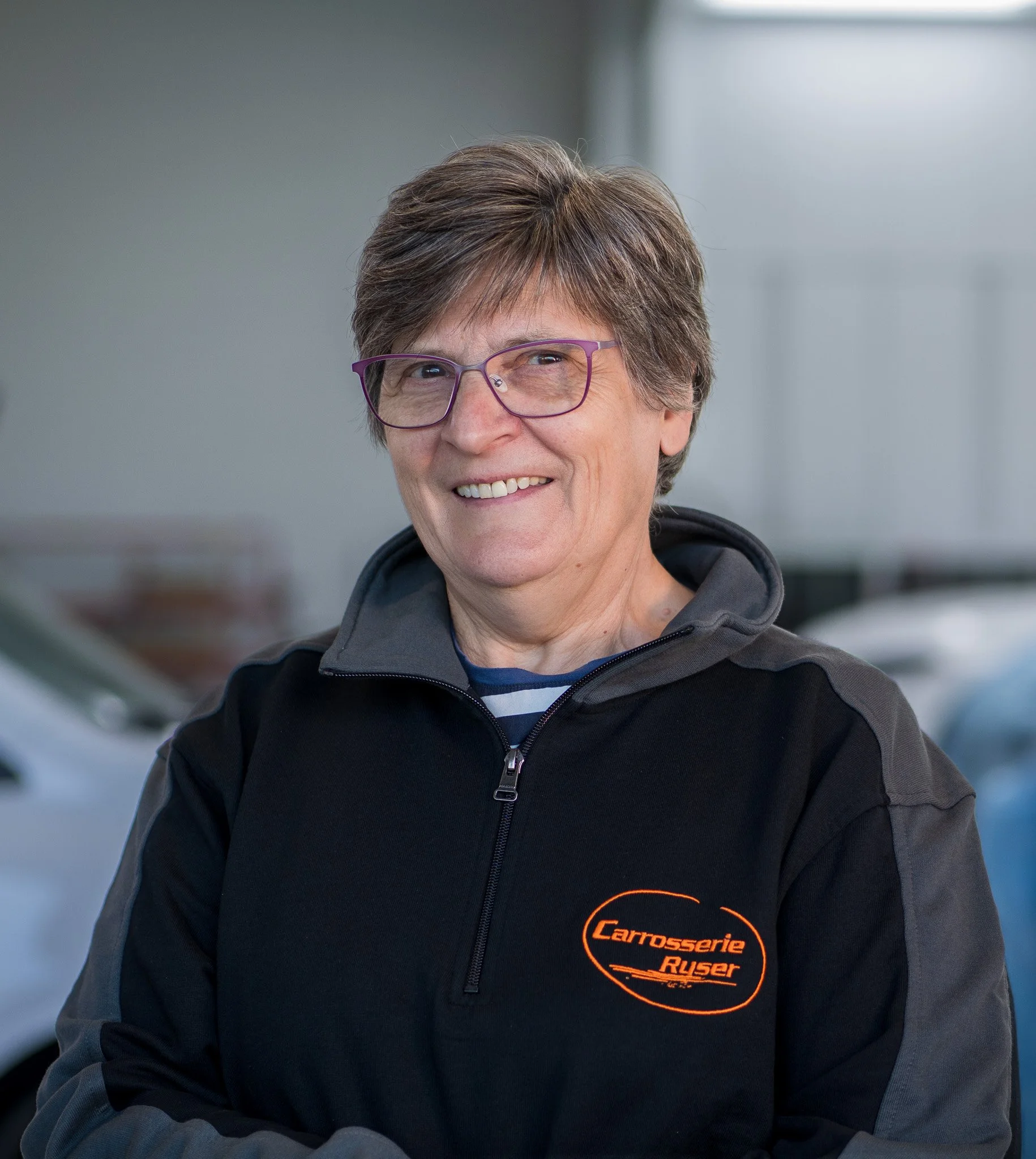 Nahaufnahme eines lächelnden älteren Mannes mit grauen Haaren und Brille, trägt eine schwarze Jacke mit einem Logo von Carrosserie Ruser in orange.