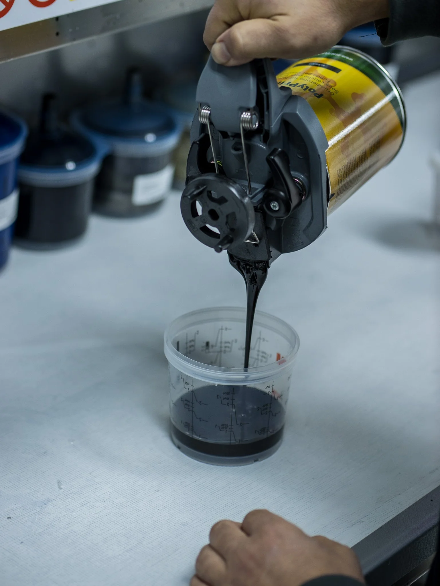 Hand hält eine Drucknadel über ein Behälter mit schwarzer Tinte auf einem Tisch, im Hintergrund sind kleine Dosen sichtbar.