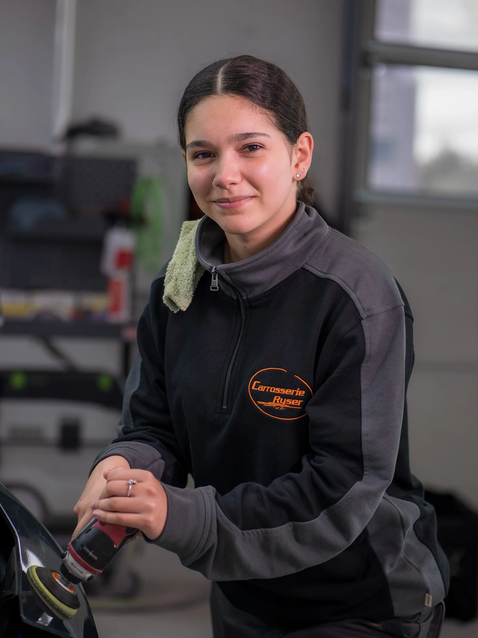 Junge Frau beim Polieren eines Autos im Werkstattraum, trägt einen schwarzen Pullover mit einem Logo, kurze braune Haare, lächelt leicht, im Hintergrund Werkzeuge und eine Fenster.