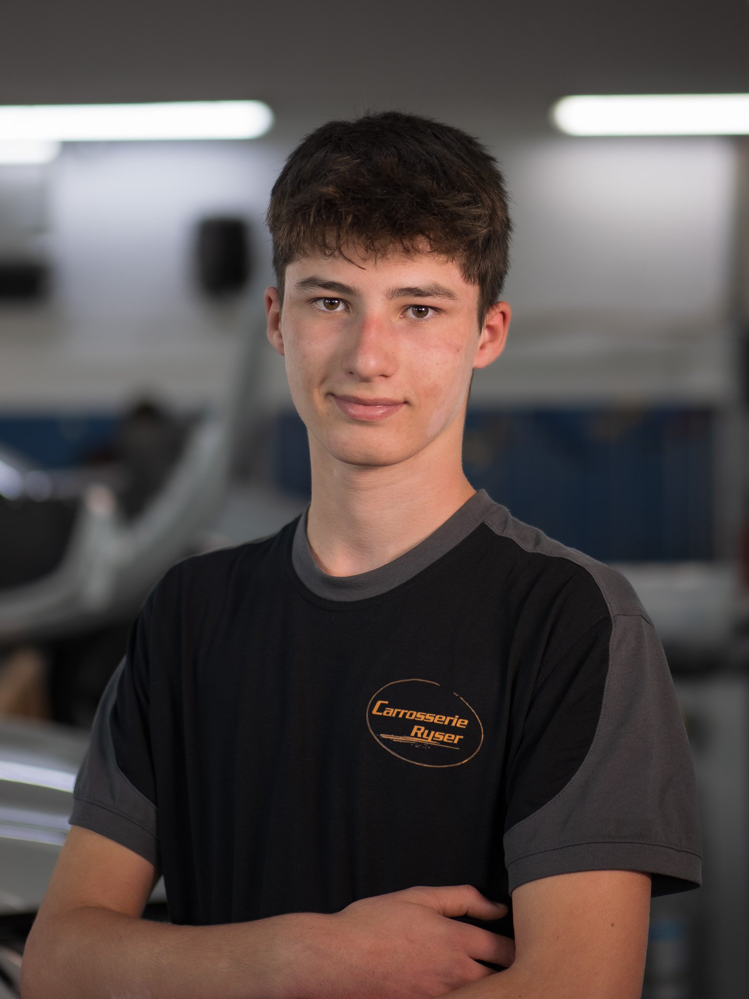 Junger Mann mit dunklen Haaren, trägt ein schwarzes T-Shirt mit Logo 'Carrosserie Ryser', steht vor einer Werkstatt, mit verschmutztem Gesicht, lächelt leicht.