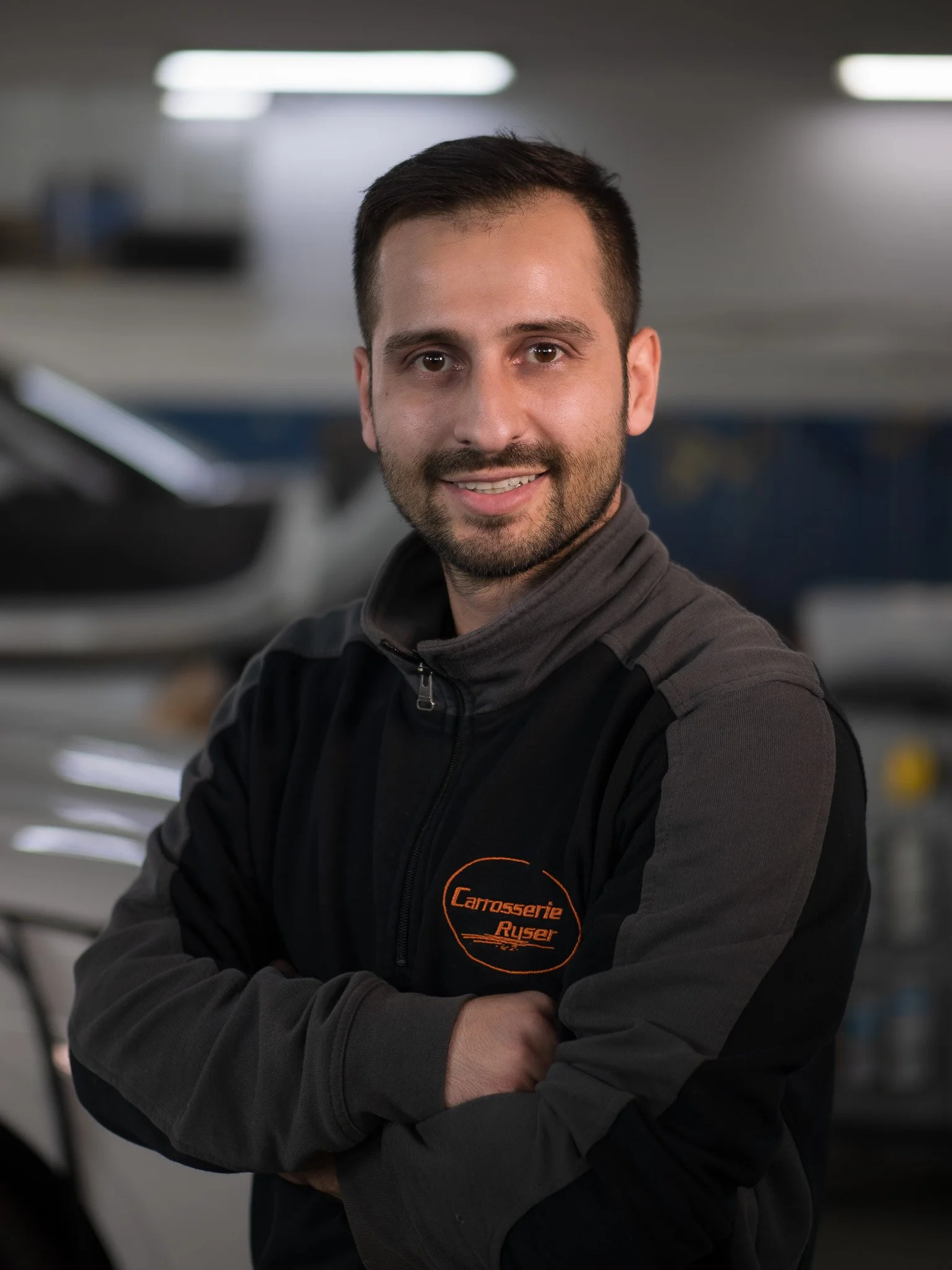 Junger Mann mit kurzen dunklen Haaren und Bart, trägt eine schwarze Jacke mit einem orangefarbenen Logo, steht mit verschränkten Armen in einer Werkstatt, hinter ihm sind Autos und Werkbank sichtbar.