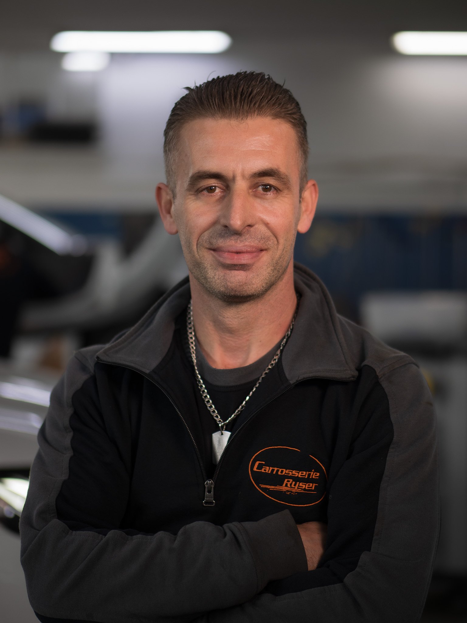 Mann mit kurzen braunen Haaren und schwarzen Jacke, stehend mit verschränkten Armen, im Hintergrund eine Werkstatt mit Autos.