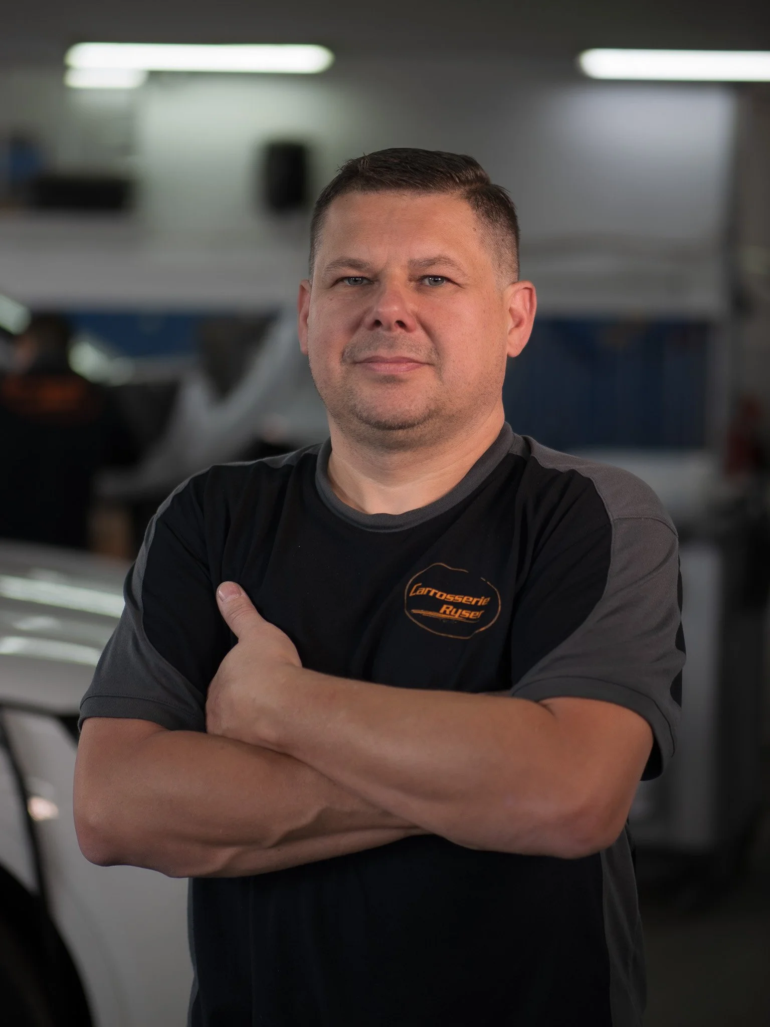 Mann in schwarzem T-Shirt mit Aufdruck 'Carrosserie Rüser' in einer Werkstatt mit seinen Armen verschränkt.