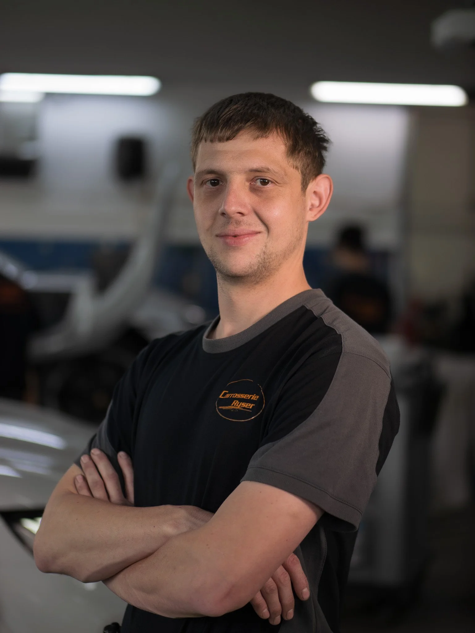 Ein junger Mann mit kurzen braunen Haaren, trägt ein schwarzes T-Shirt mit einem Logo 'Carrosserie Ruser', steht vor einem blassen Hintergrund in einer Werkstatt und schaut in die Kamera.