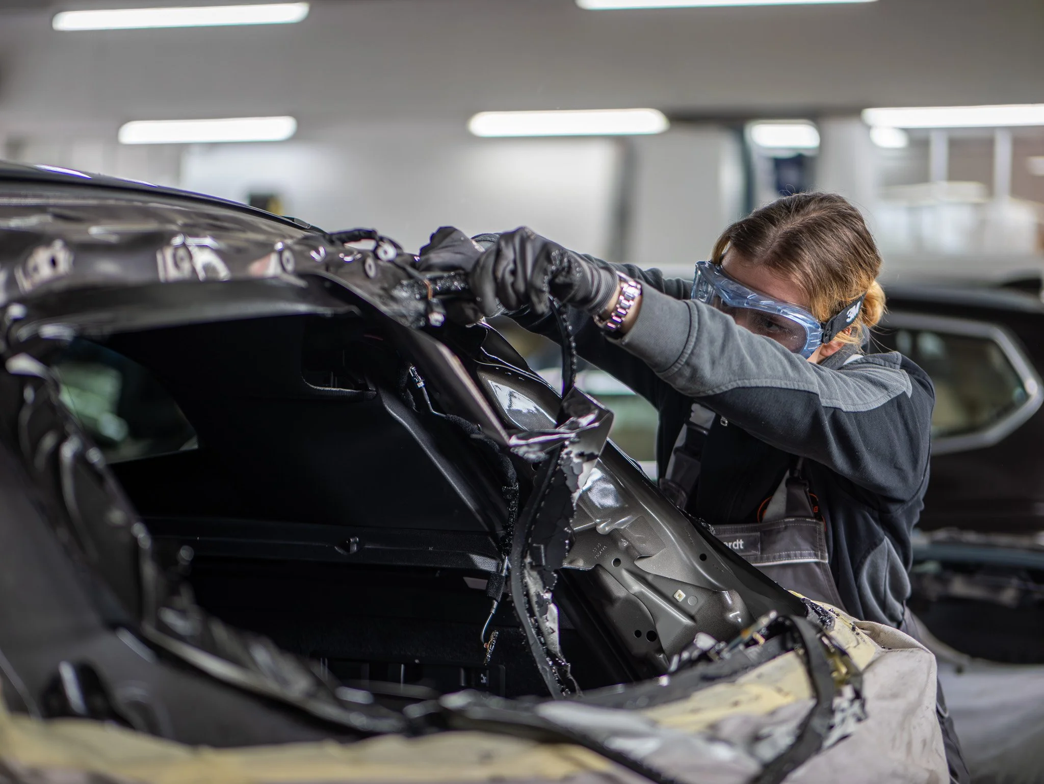 Eine Carosserie Spenglerin arbeitet an einem schwarzen Auto im Industriegebiet, trägt Schutzbrille, schwarze Handschuhe, und einen grauen Anzug.