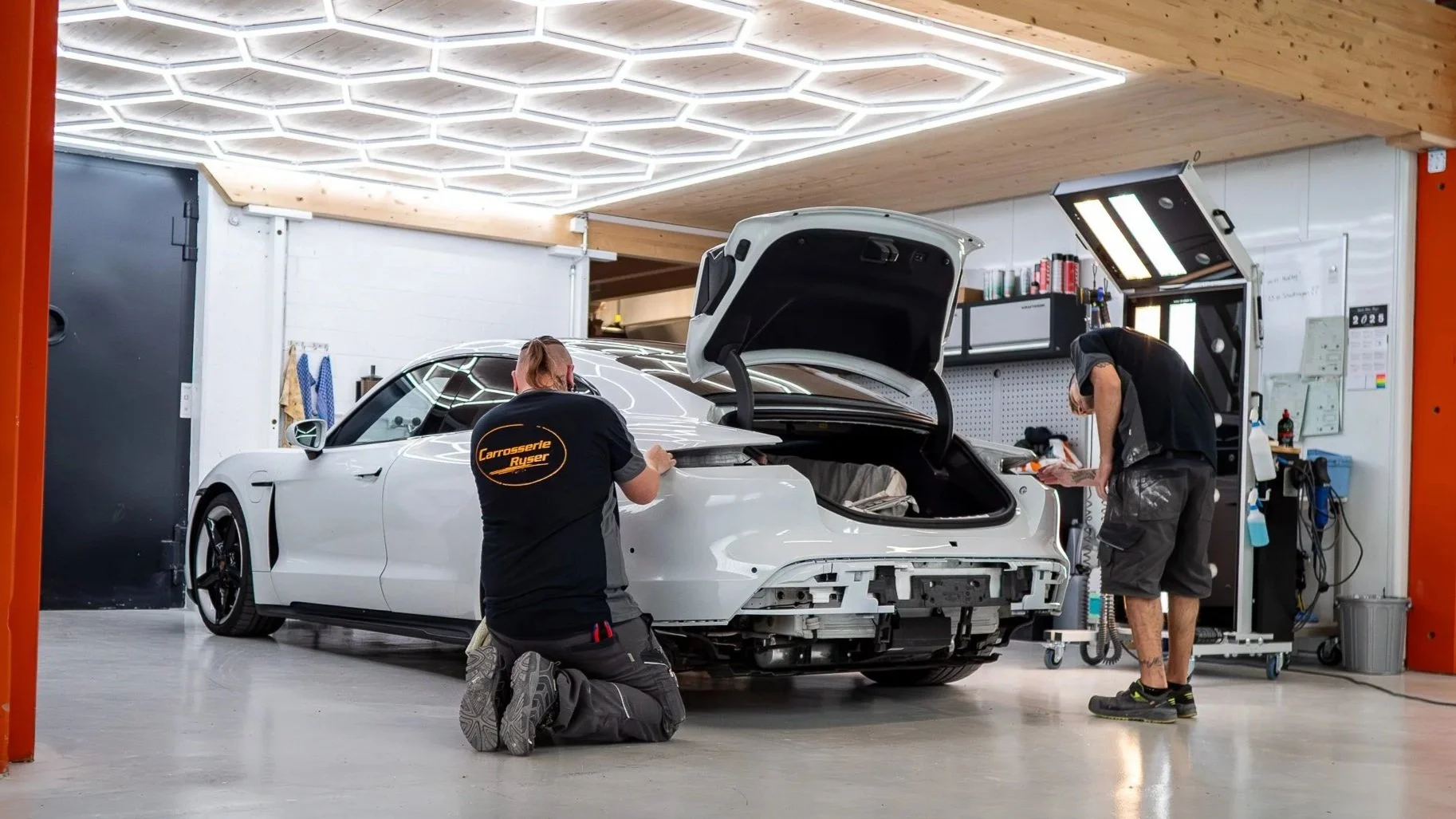 Zwei Mechaniker arbeiten an einem weissen Sportwagen in einer Werkstatt, einer ist kniend und der andere steht, beide tragen schwarze Arbeitskleidung.