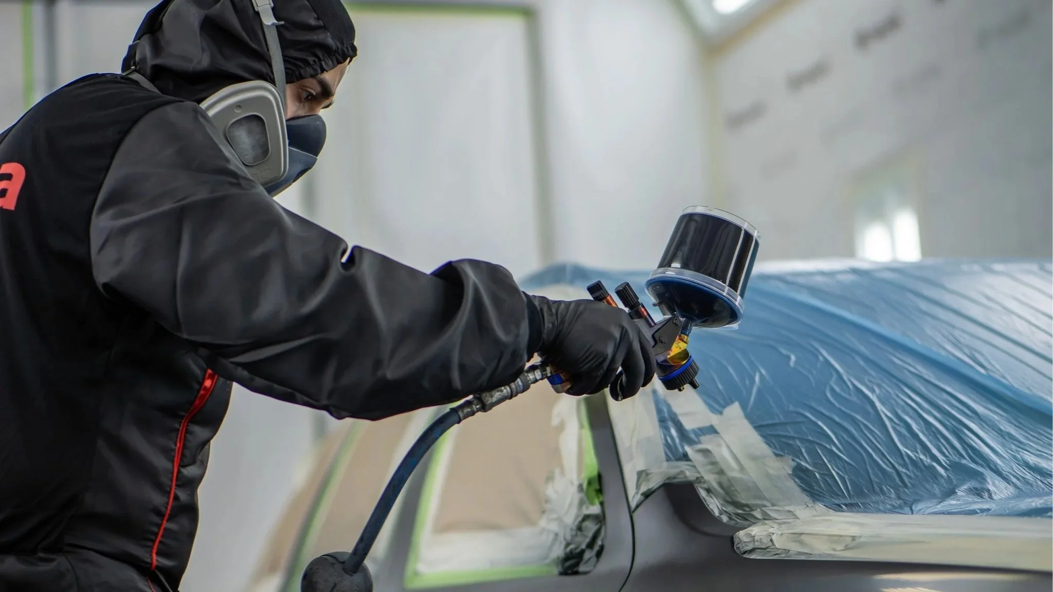 Ein Techniker trägt Schutzkleidung und maskiert und sprayt Autolack in einer Werkstatt.