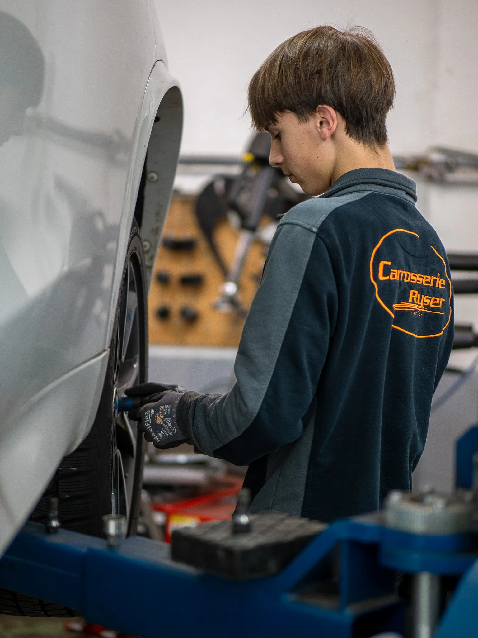 Ein junger Mann arbeitet in einer Autowerkstatt an einem Auto. Er trägt eine Arbeitsjacke mit dem Logo "Campsserie Rüser" auf dem Rücken.