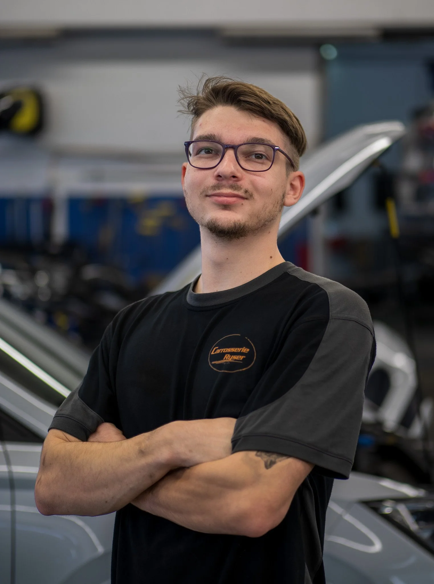 Ein junger Mann mit Brille, kurzen braunen Haaren und einem leichten Bart, der in einem Werkstatt oder Garage steht. Er trägt ein schwarzes T-Shirt mit einem Logo, das 'Carrosserie Rytzer' heißt, und hat seine Arme verschränkt.