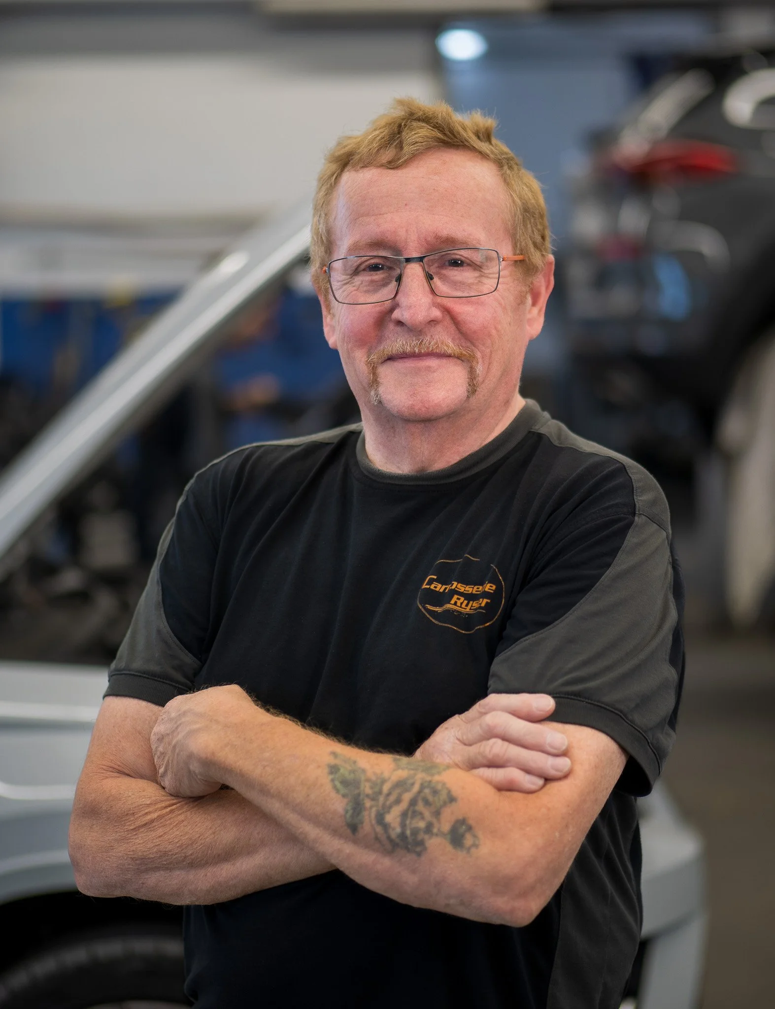 Mann mit Brille, blondem Haar und Schnäuzer, der lächelt und die Arme verschränkt, trägt ein schwarzes T-Shirt mit Logo, im Hintergrund eine Werkstatt mit Auto.