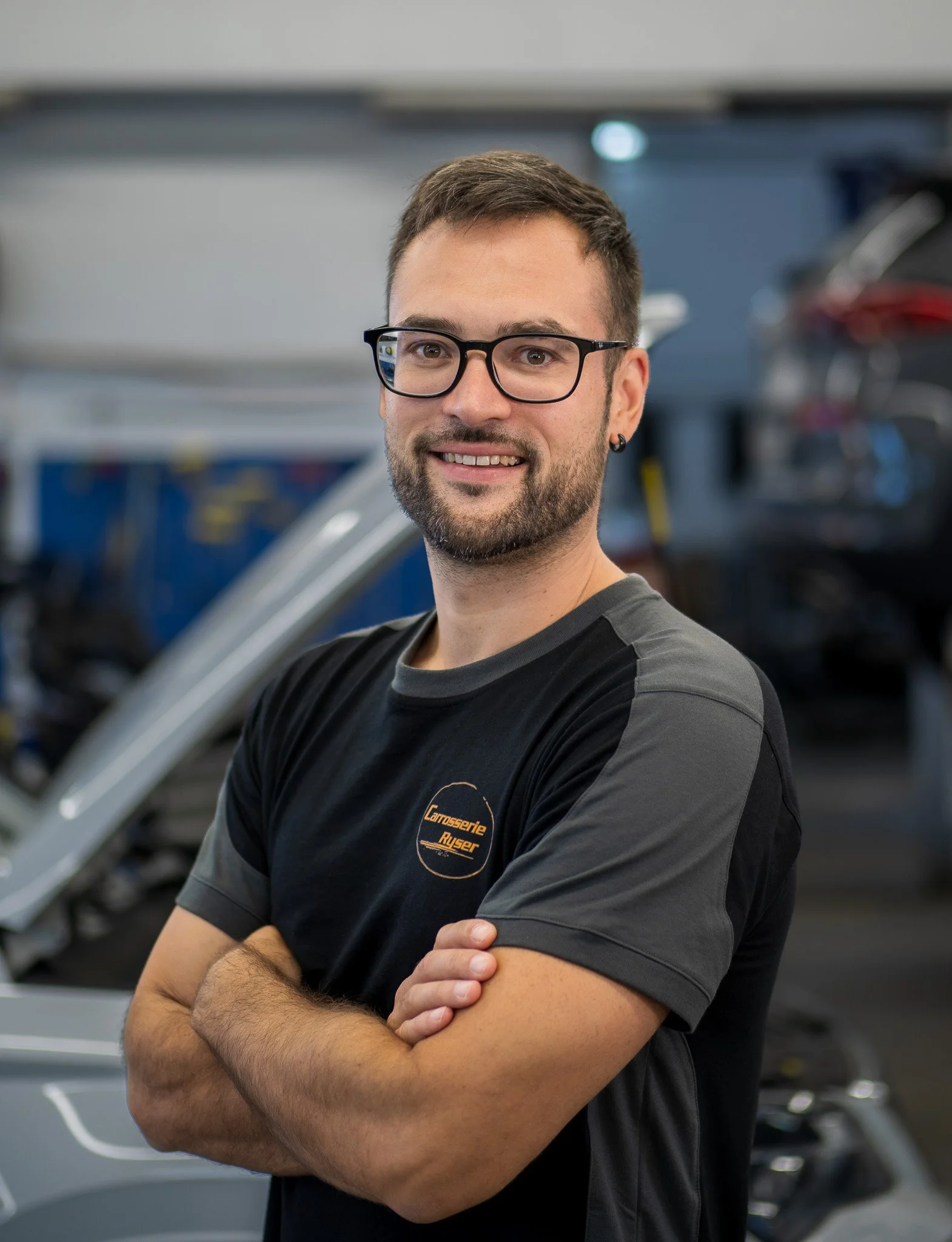 Ein Mann mit Brille und Bart steht in einer Werkstatt, trägt ein schwarzes T-Shirt mit einem Logo der Carrosserie Ruser, und hat die Arme verschränkt.