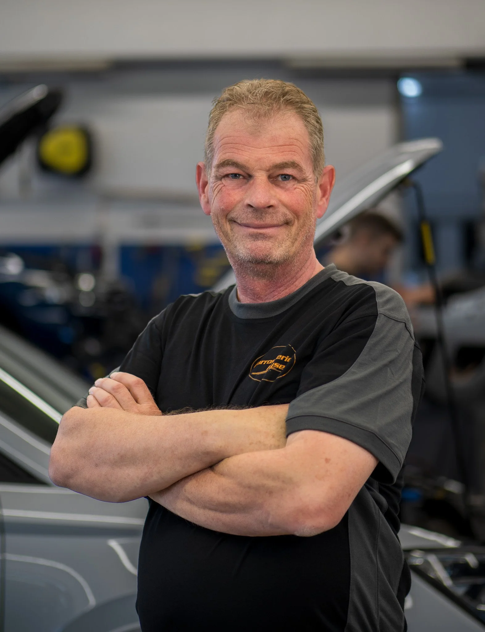 Ein älterer Mann mit kurzen, lockigen, hellbraunen Haaren und einem freundlichen Lächeln, der eine schwarze T-Shirt trägt, steht vor einer Garage mit mehreren Fahrrädern im Hintergrund.