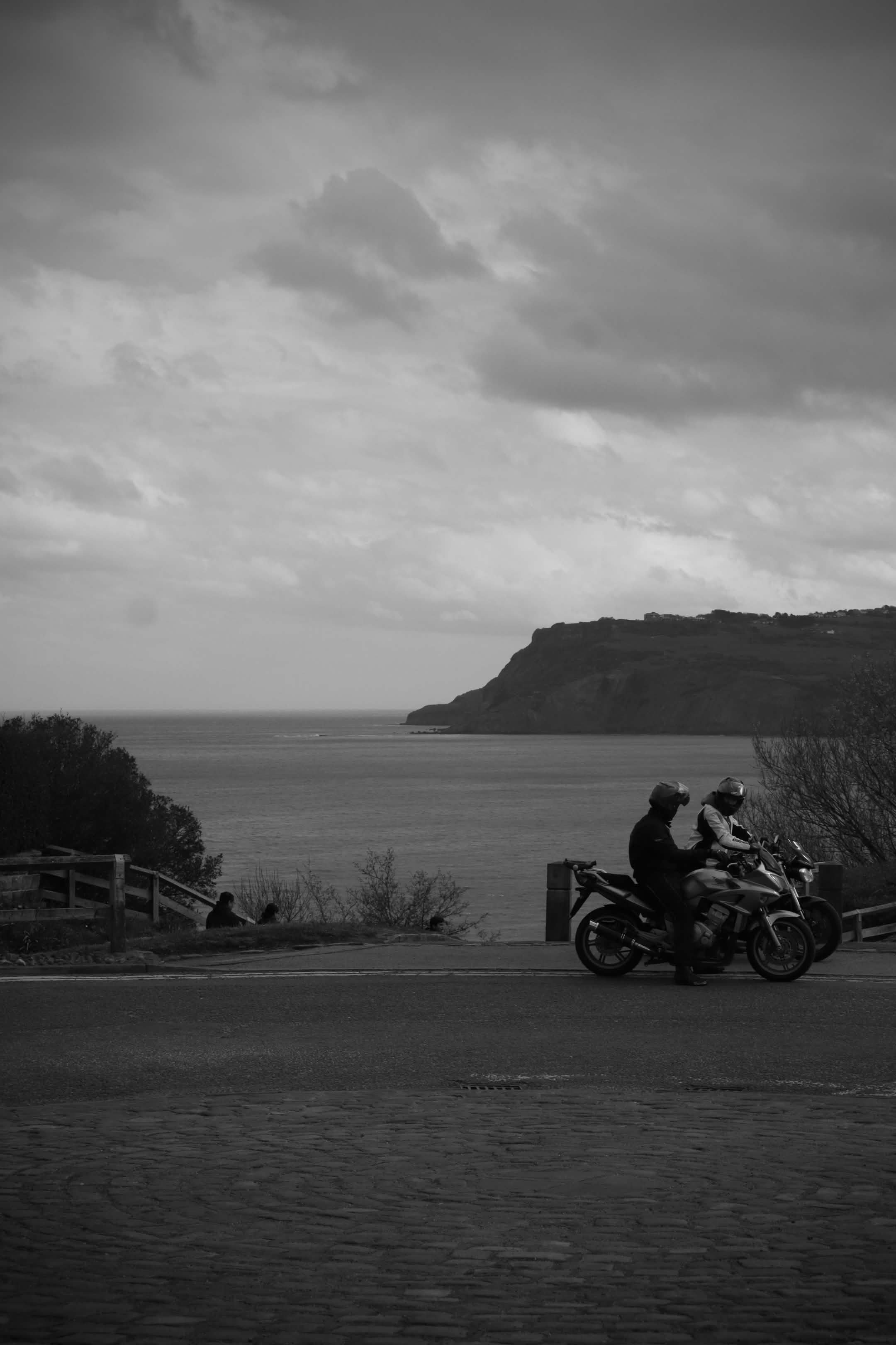 Black and white photo of two people riding a motorcycle along a coastal road with a cliff, water, and cloudy sky in the background.