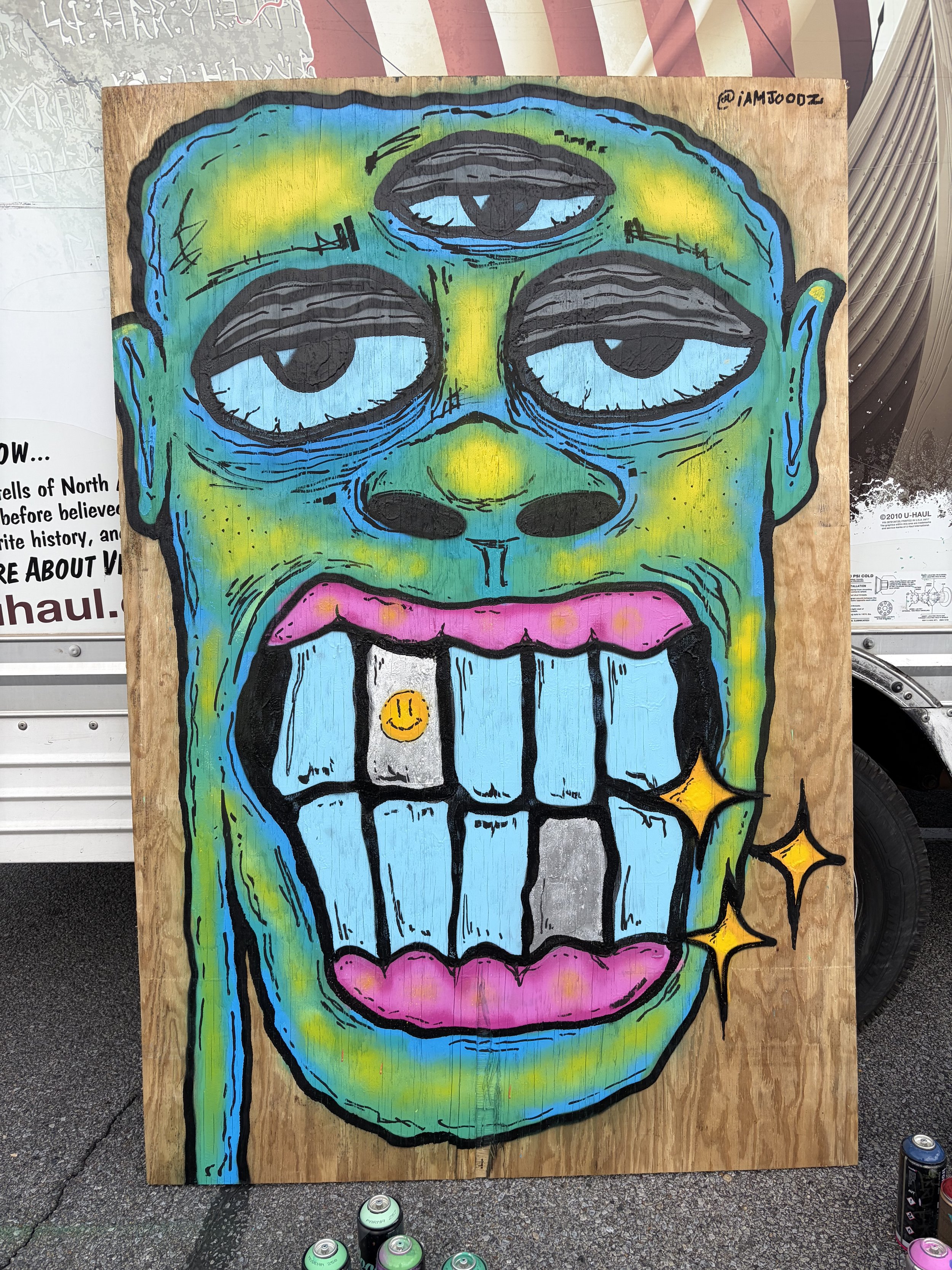 Colorful three-eyed character painting on wood by IAMJOODZ featuring blue, green, yellow and pink details.