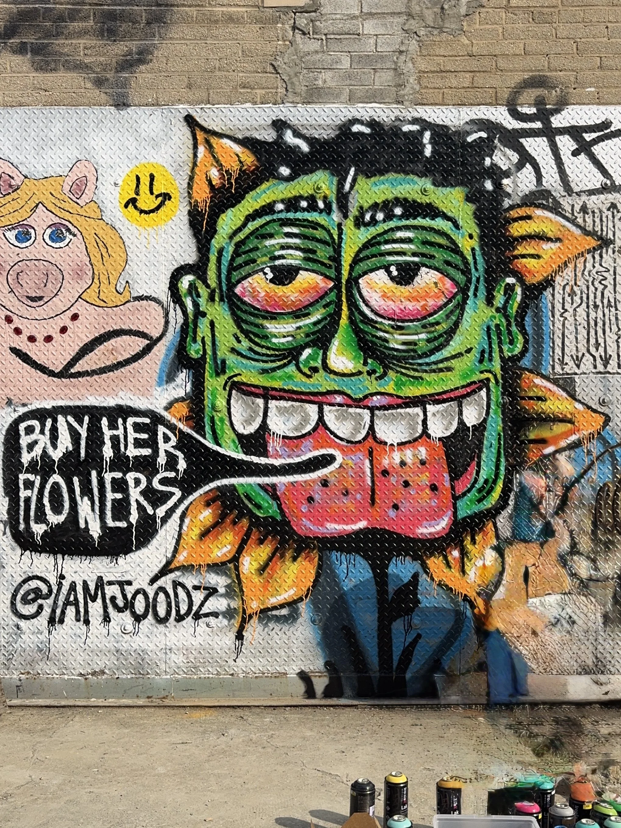 Buy Her Flowers street art mural by IAMJOODZ featuring a green character surrounded by orange flowers.