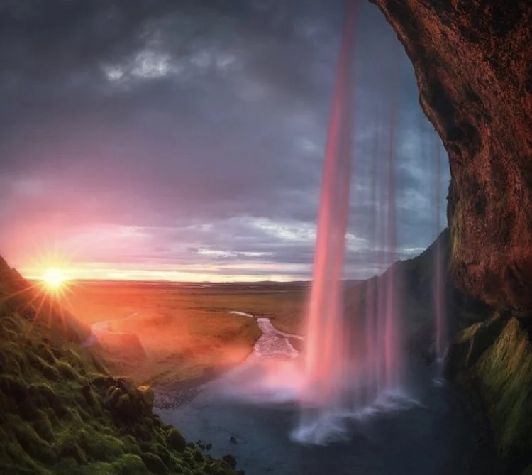 Scenic view of a waterfall with pinkish hue, a river, and a sunset in the distance under cloudy skies.