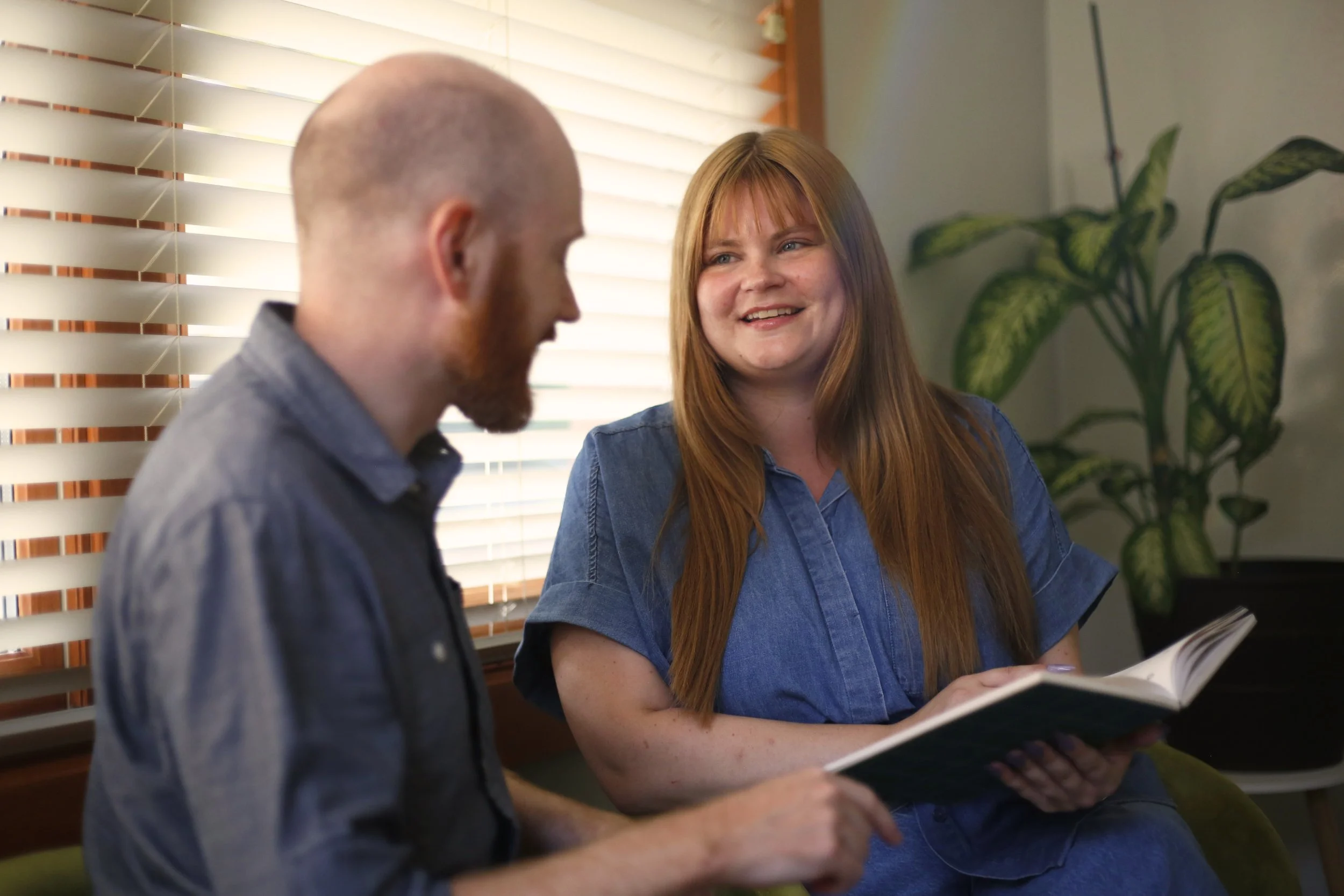 A man with a bald head and a woman with long red hair smiling and talking while looking at a book indoors.