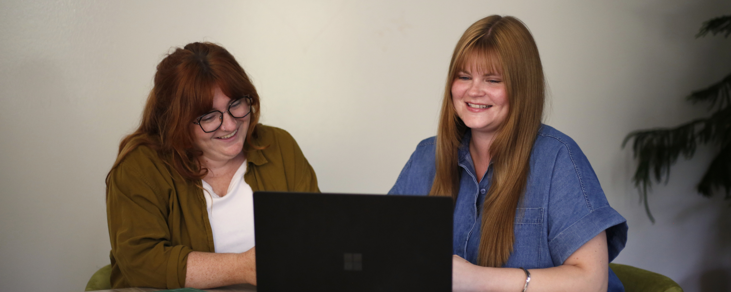Two women sitting at a table looking at a laptop and smiling.