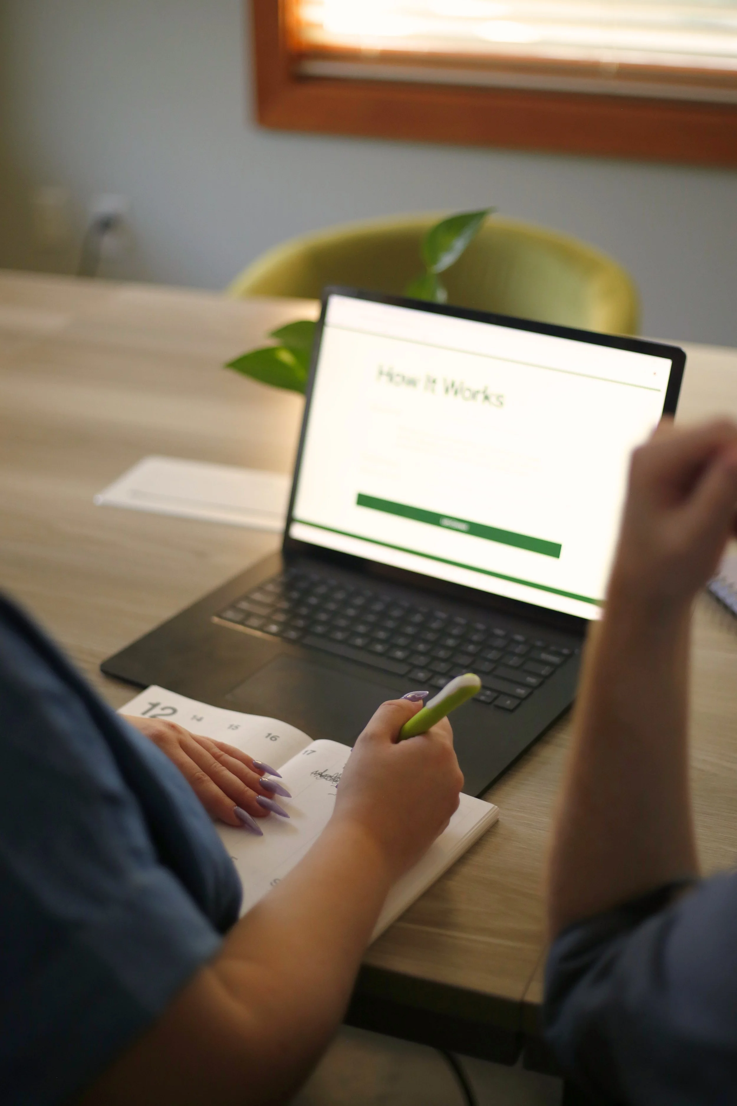 Person working on a laptop with a presentation slide titled 'How it Works' visible on the screen. They are also writing in a planner or notebook with handwritten notes. The scene takes place at a desk near a window with blinds, with a yellow chair and a plant in the background.