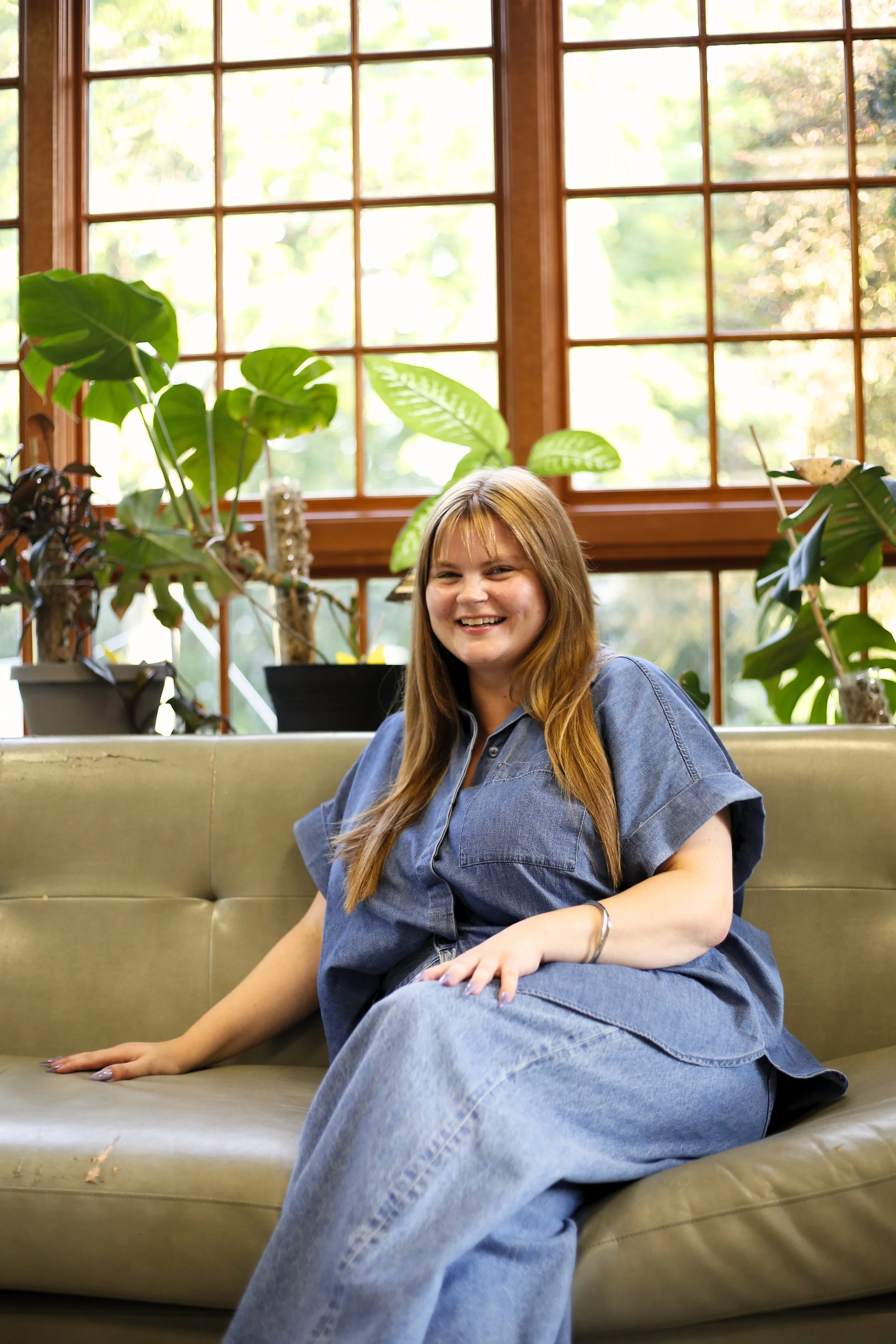 A young woman with long light brown hair, wearing a denim shirt and jeans, sitting on a beige couch in a room with large window panes and green plants, smiling at the camera.