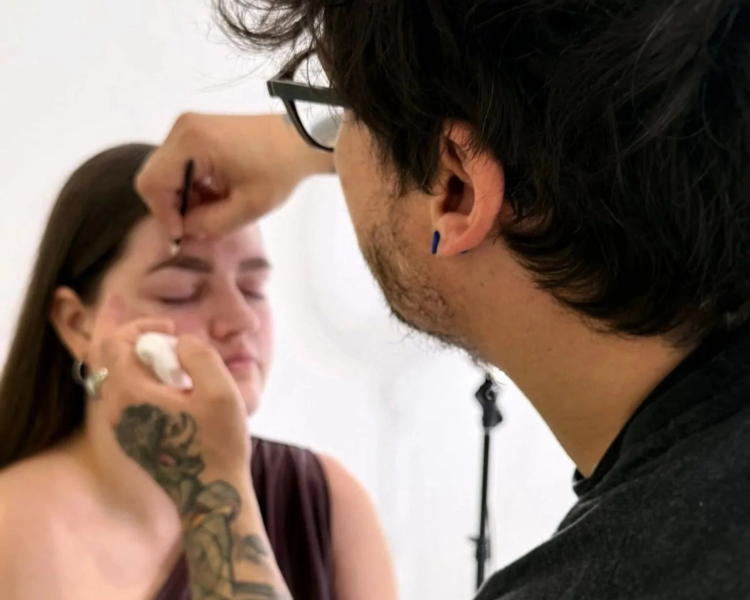 Un homme tatoué maquille une femme avec une brosse à maquillage sur le visage.