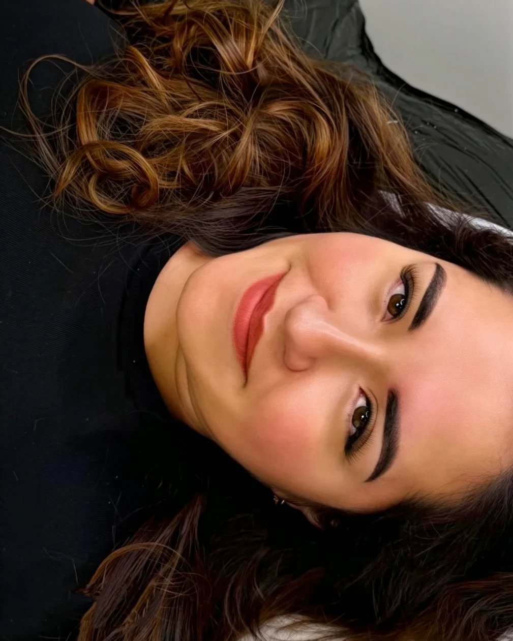 Une femme avec de longs cheveux bouclés, allongée sur un lit, souriant à la caméra avec des sourcils fraîchement tatoués.