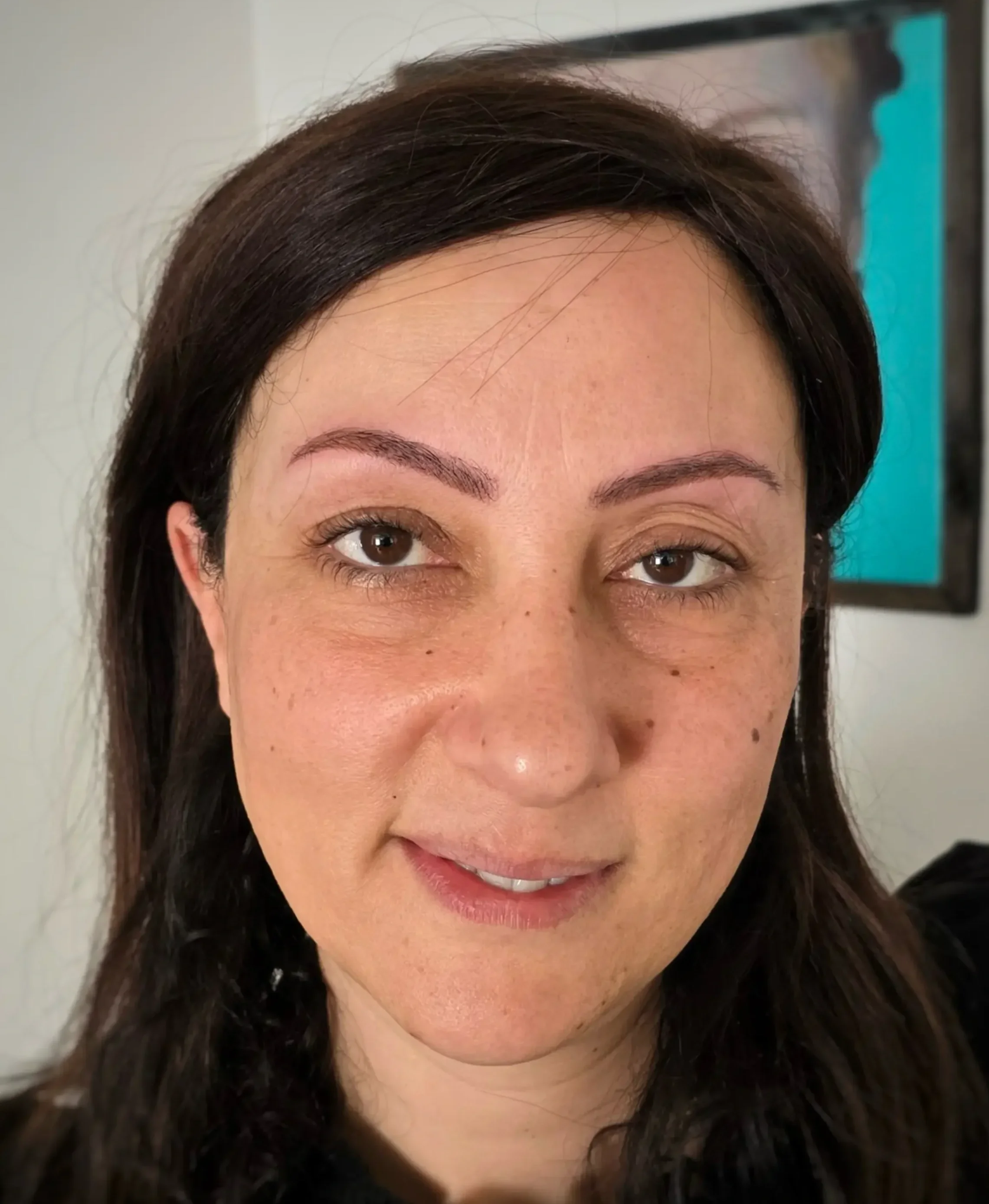 Photo d'une femme avec des cheveux bruns foncés, souriante, prise de près portant un microblading tout neuf. Background flou.