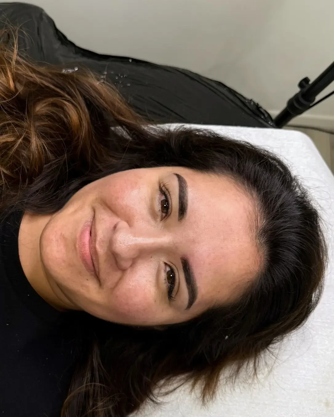 Une femme allongée sur un lit, souriante, avec des cheveux bruns et des sourcils bien dessinés, portant un haut noir.