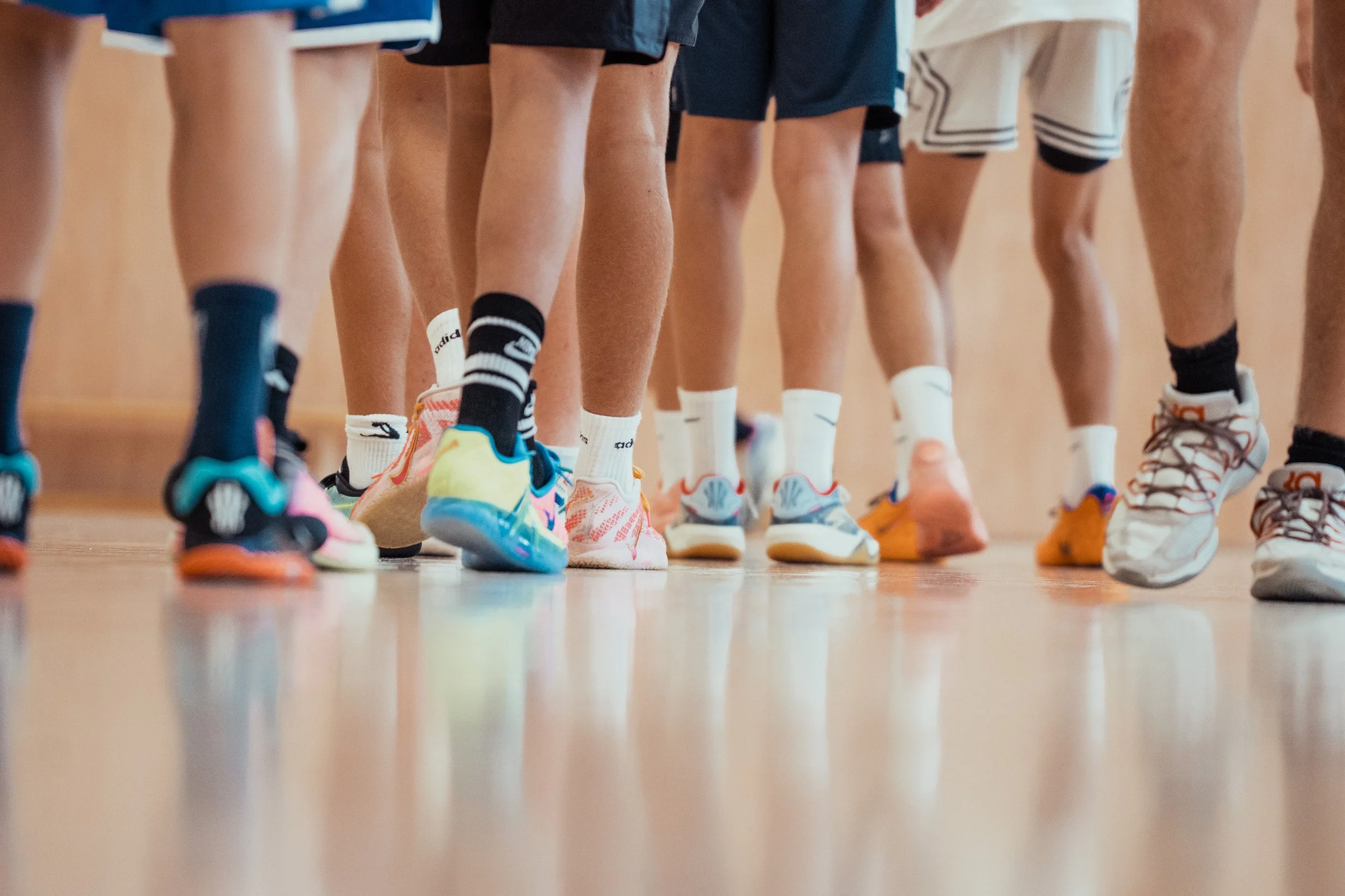 Close-up view of multiple children's legs and feet in athletic shoes and socks standing in a line on a gymnasium floor.