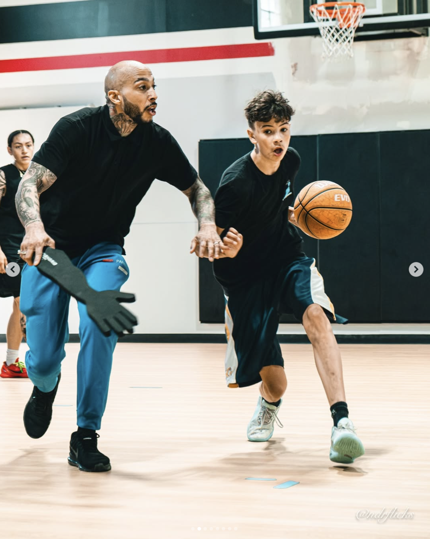 A man and a boy playing basketball on an indoor court, with the man defending the boy who is dribbling the ball. There is a woman in the background.