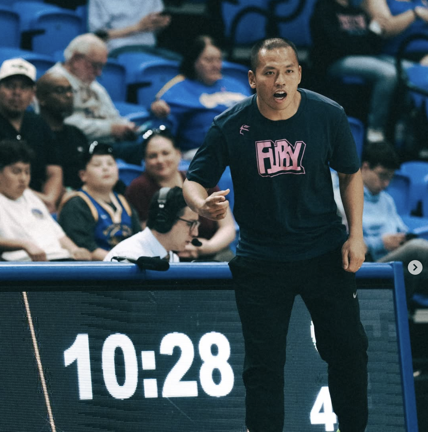 A man in a black sports shirt with pink lettering is coaching during a volleyball match. He is standing near the sideline with a digital scoreboard showing 10:28. There are spectators and a referee sitting behind him in a stadium.