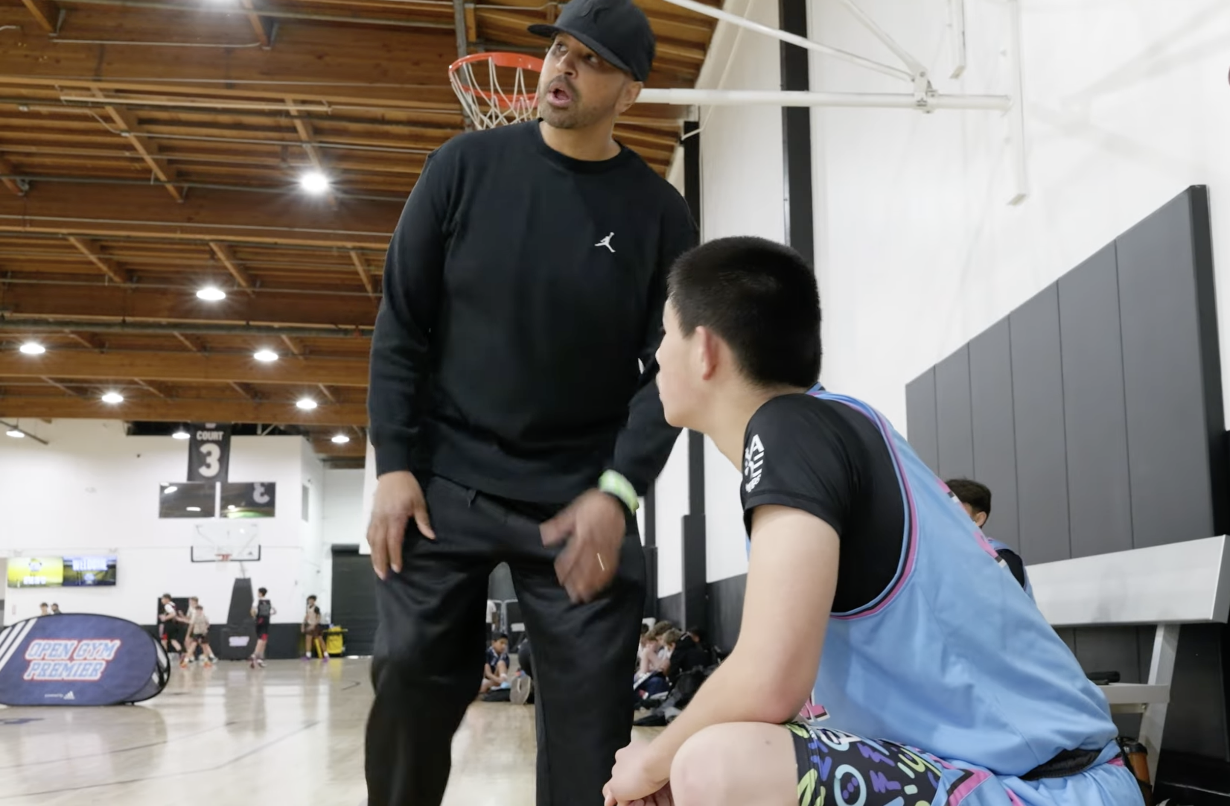 A basketball coach is standing and talking to a player, who is sitting on a bench and listening attentively. The player is wearing a black T-shirt with colorful shorts, and the coach is wearing a black hoodie and a black cap. The scene takes place in an indoor basketball court with a high wooden ceiling and basketball hoops in the background.