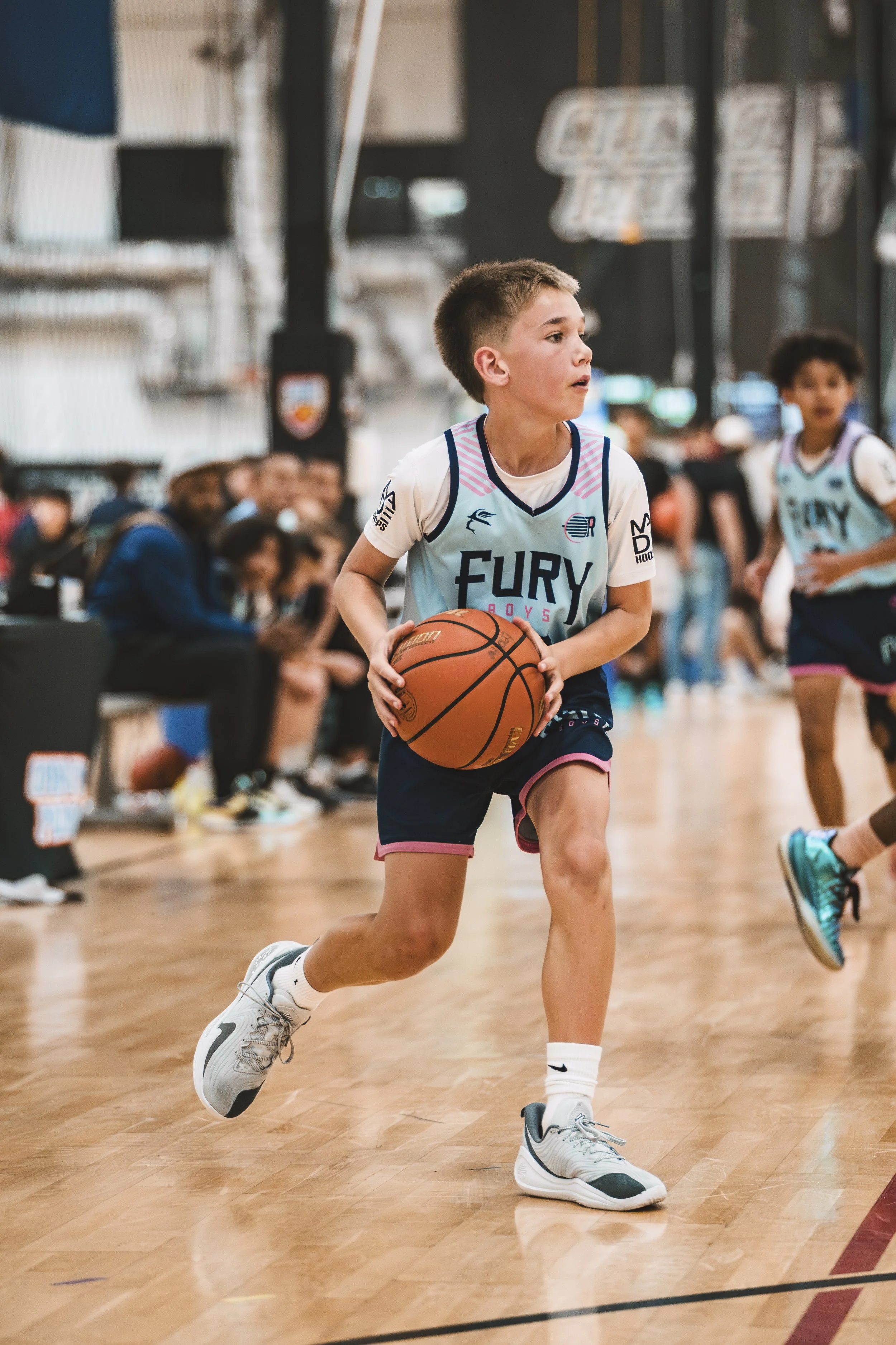 Young boy in a basketball jersey dribbling the ball on an indoor court, with spectators in the background.