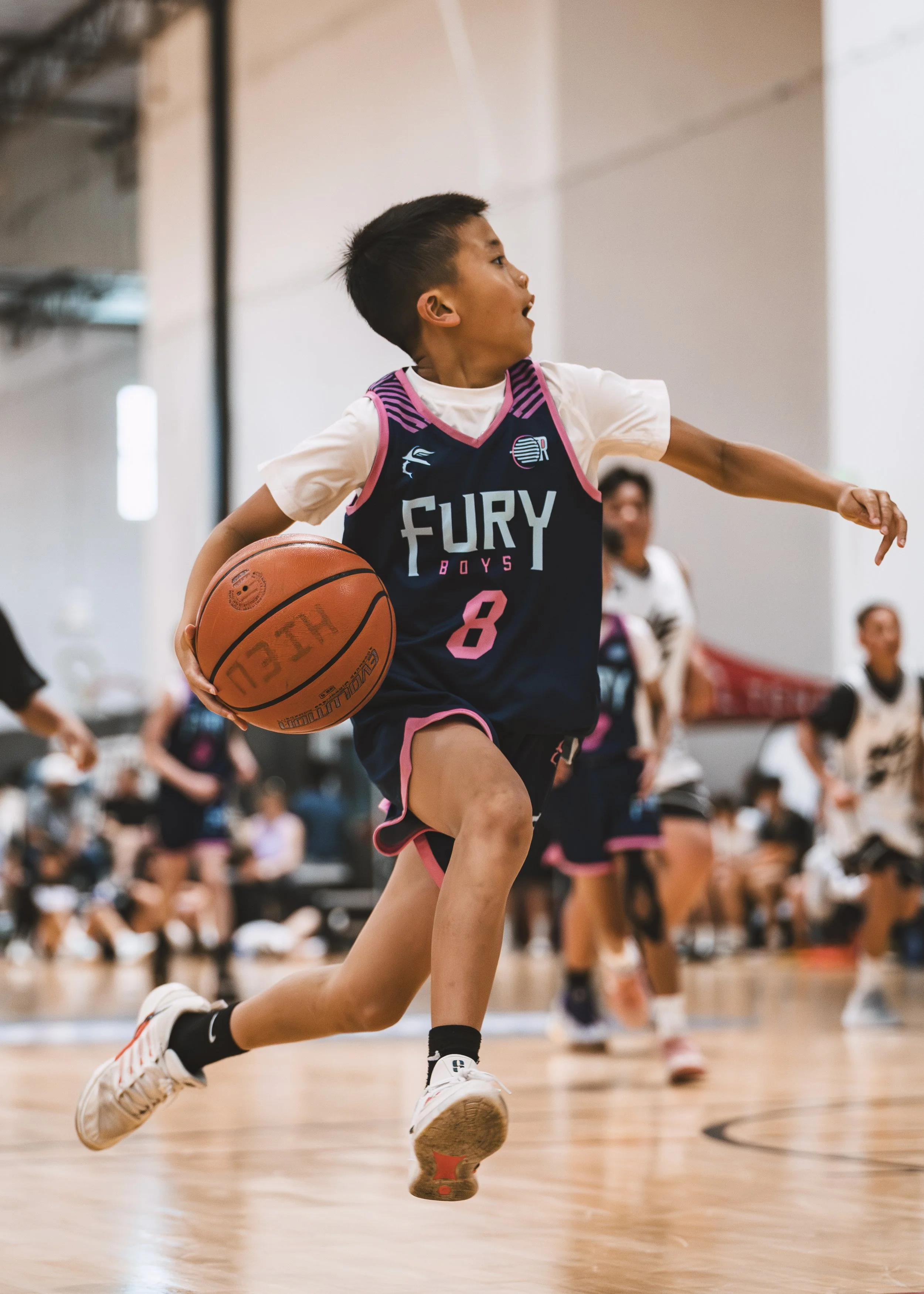 A young boy playing basketball indoors, wearing a black and pink 'Fury Boys' jersey with the number 8, holding a basketball, and running on a wooden court. Other players and spectators are in the background.