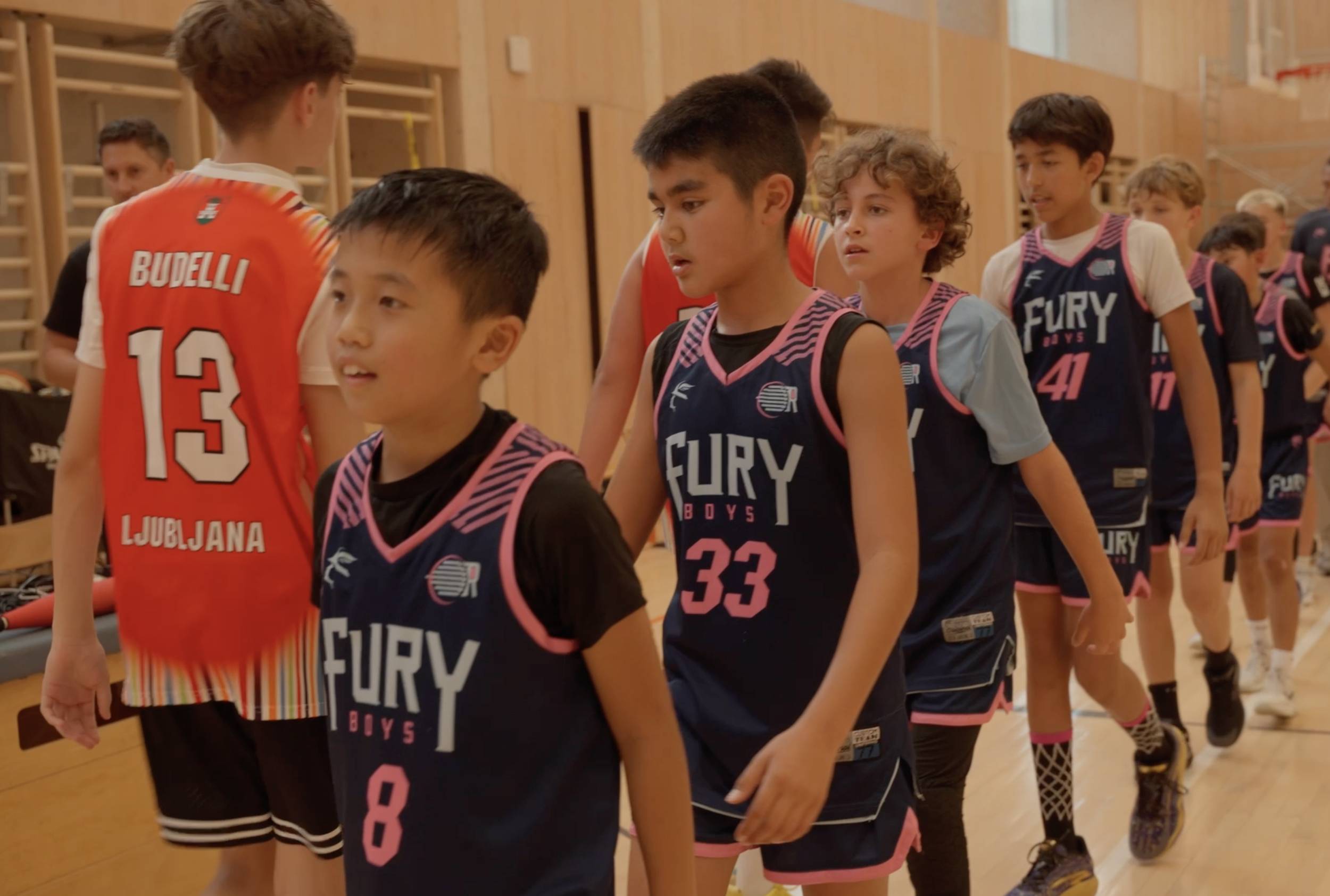 A group of young boys in basketball uniforms lined up indoors, shaking hands after a game, with a coach or referee standing in the background.