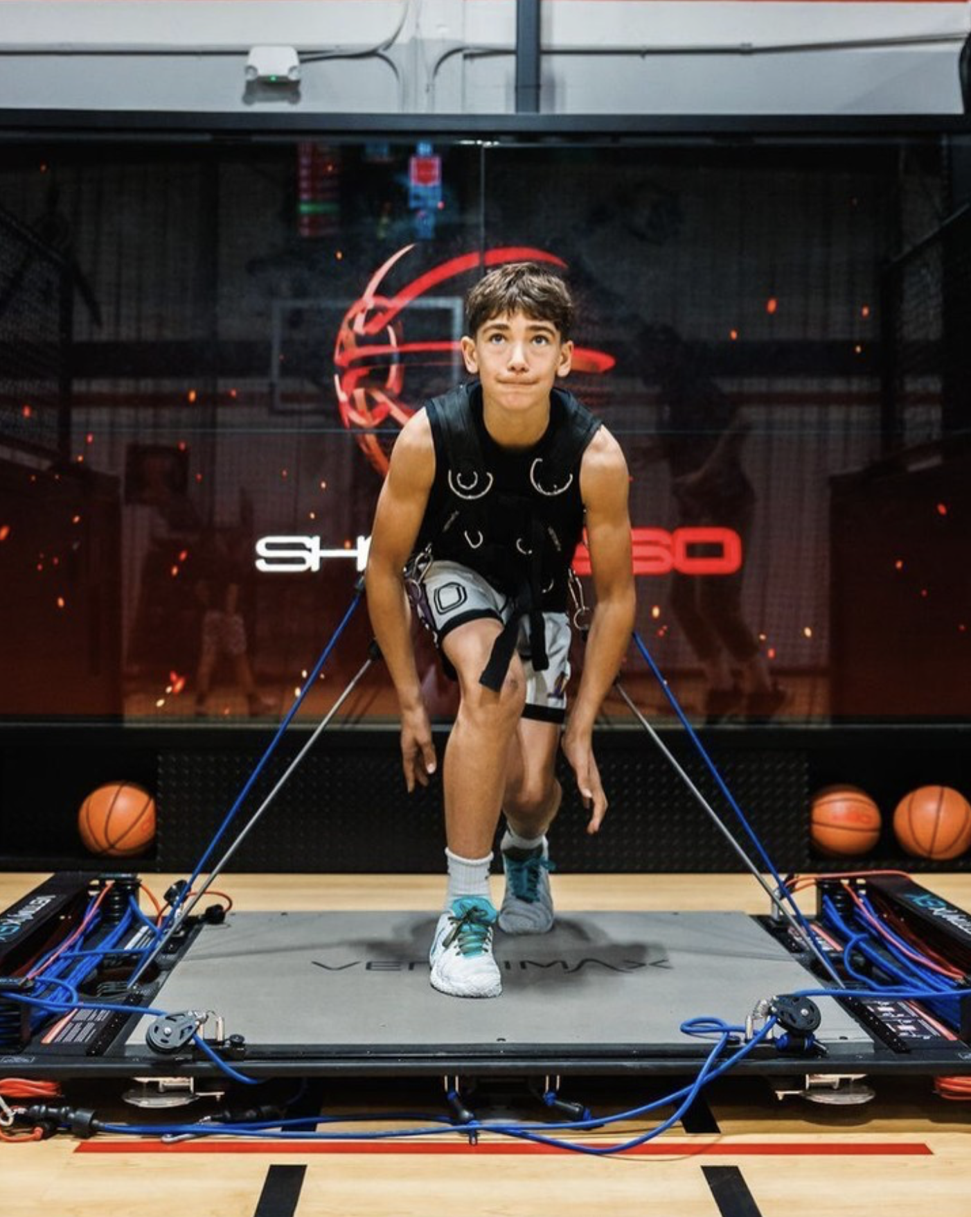 A boy running on a treadmill with sports science equipment connected to him, including wires and sensors, in a gym or sports science lab. Basketballs are on the floor beside the treadmill.