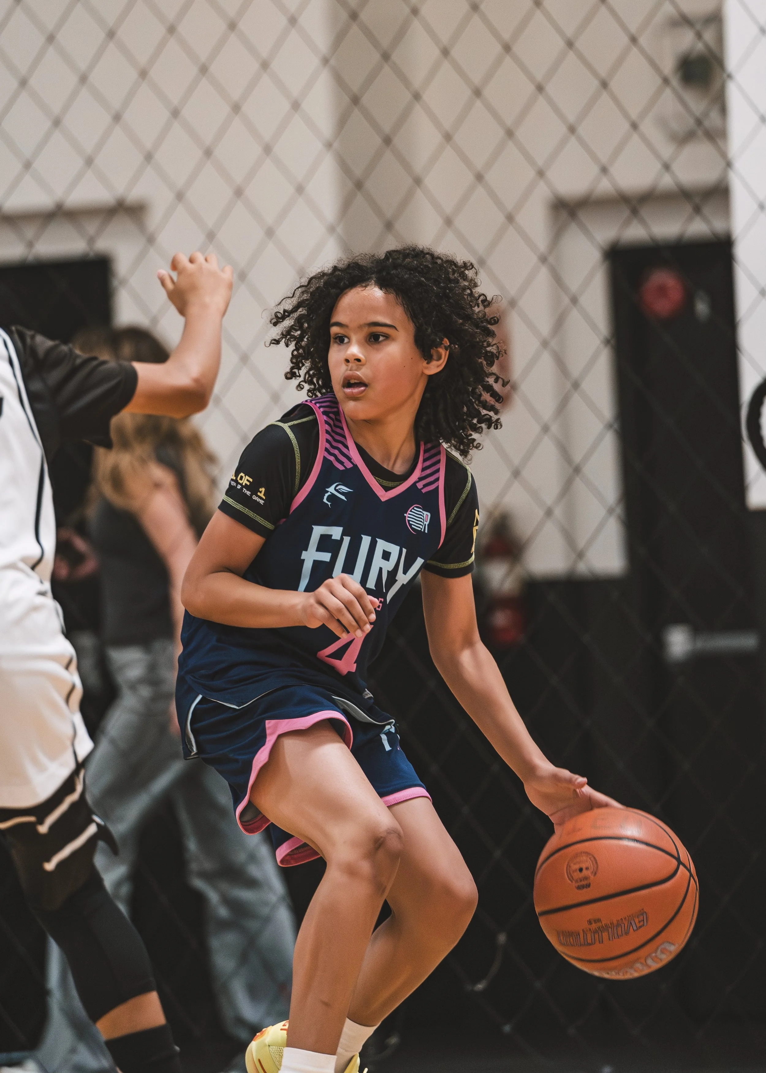 Young female basketball player with curly hair dribbling an orange basketball inside a gym, wearing a black and pink jersey, with a chain-link fence in the background.