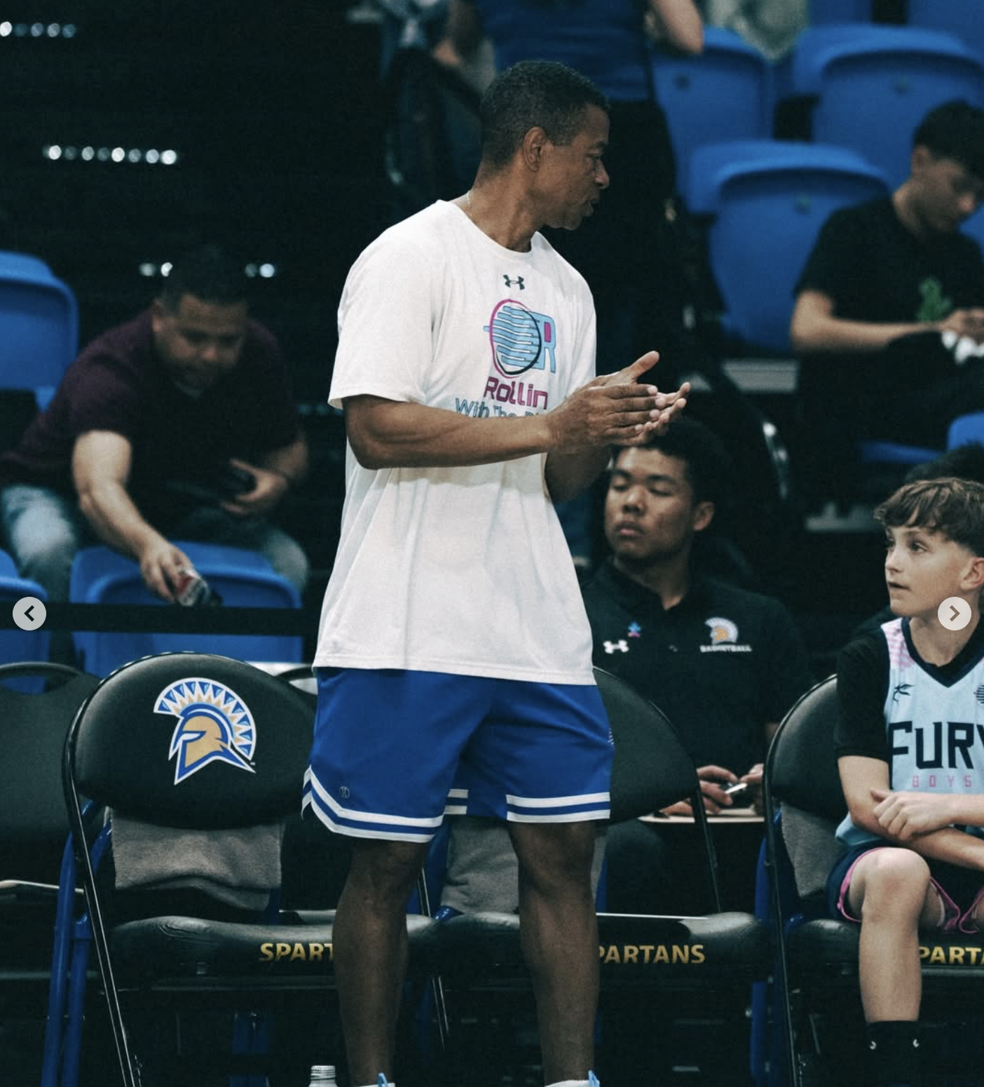 A basketball player stands on the sideline during a game, wearing a white t-shirt and blue shorts, with others seated around him in a sports arena.
