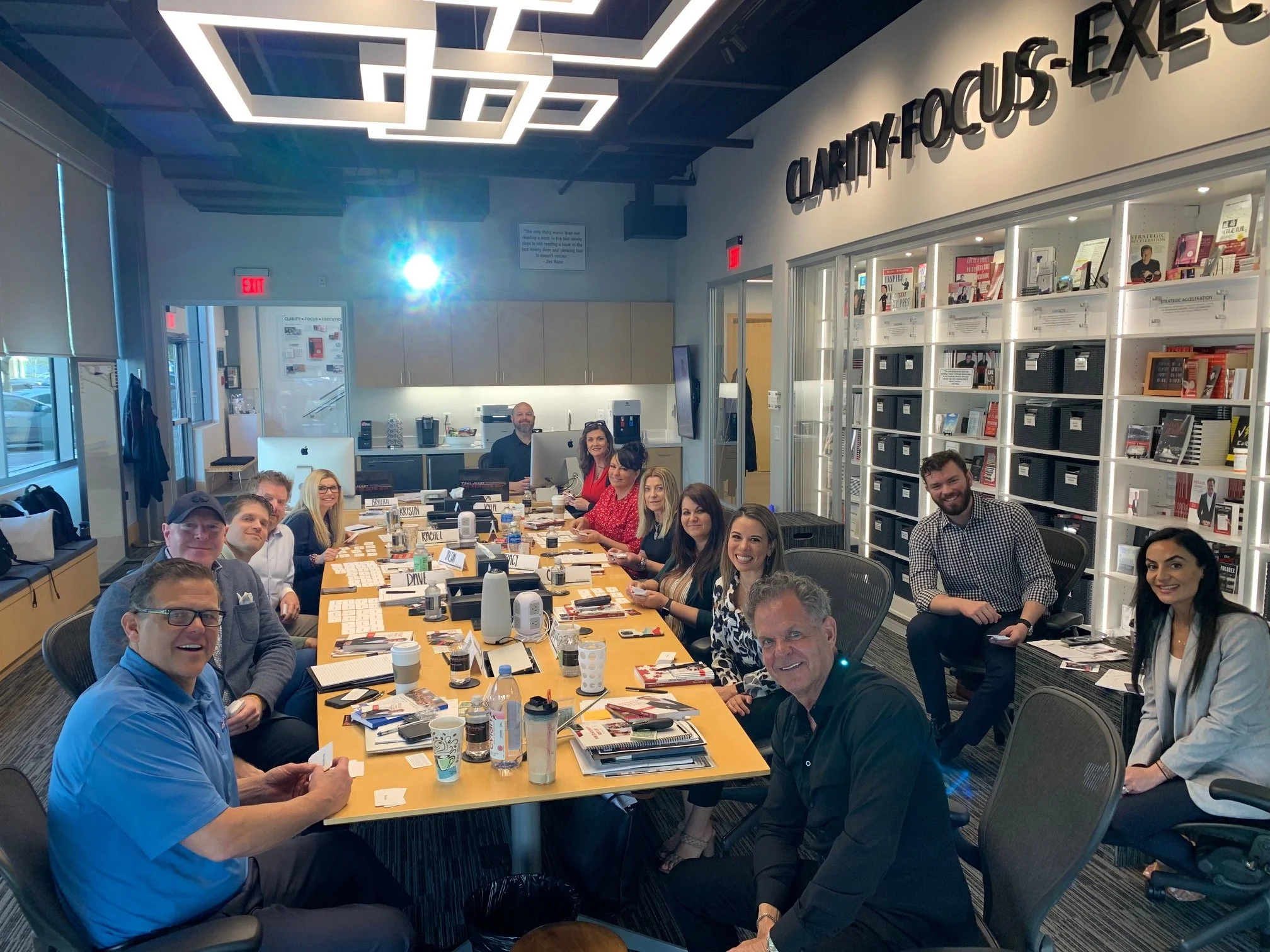 Group of smiling professionals gathered around a long conference table in a modern office, with bookshelves and decorative lighting.