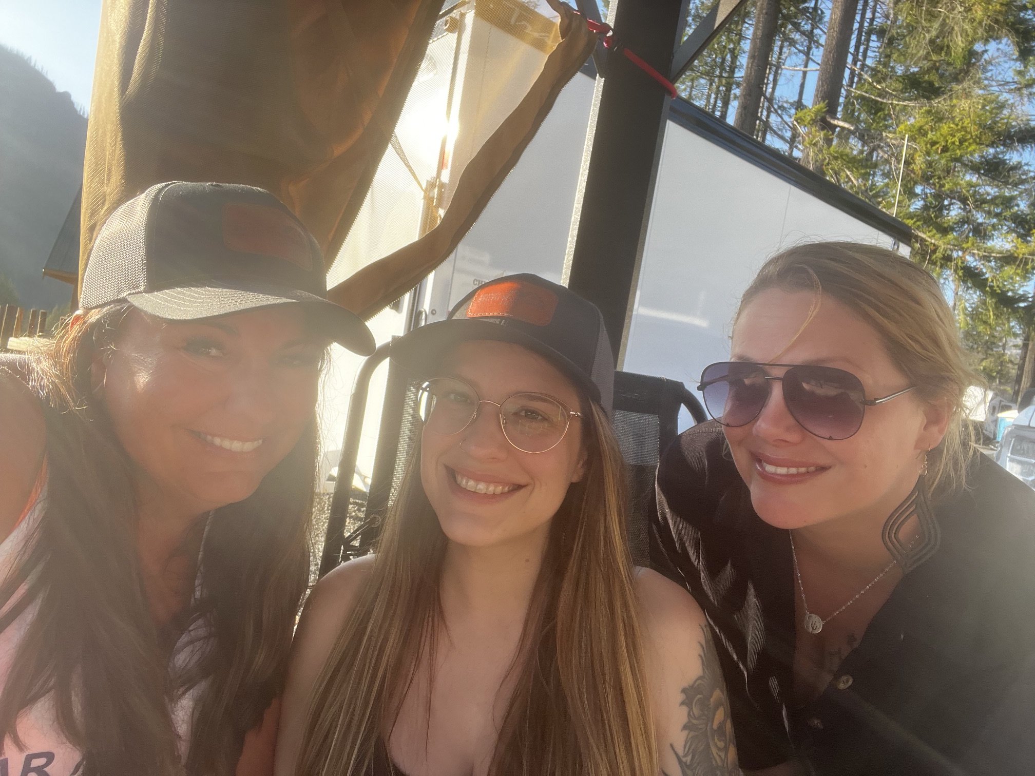Three women taking a selfie outdoors with trees in the background, wearing sunglasses and hats, smiling.