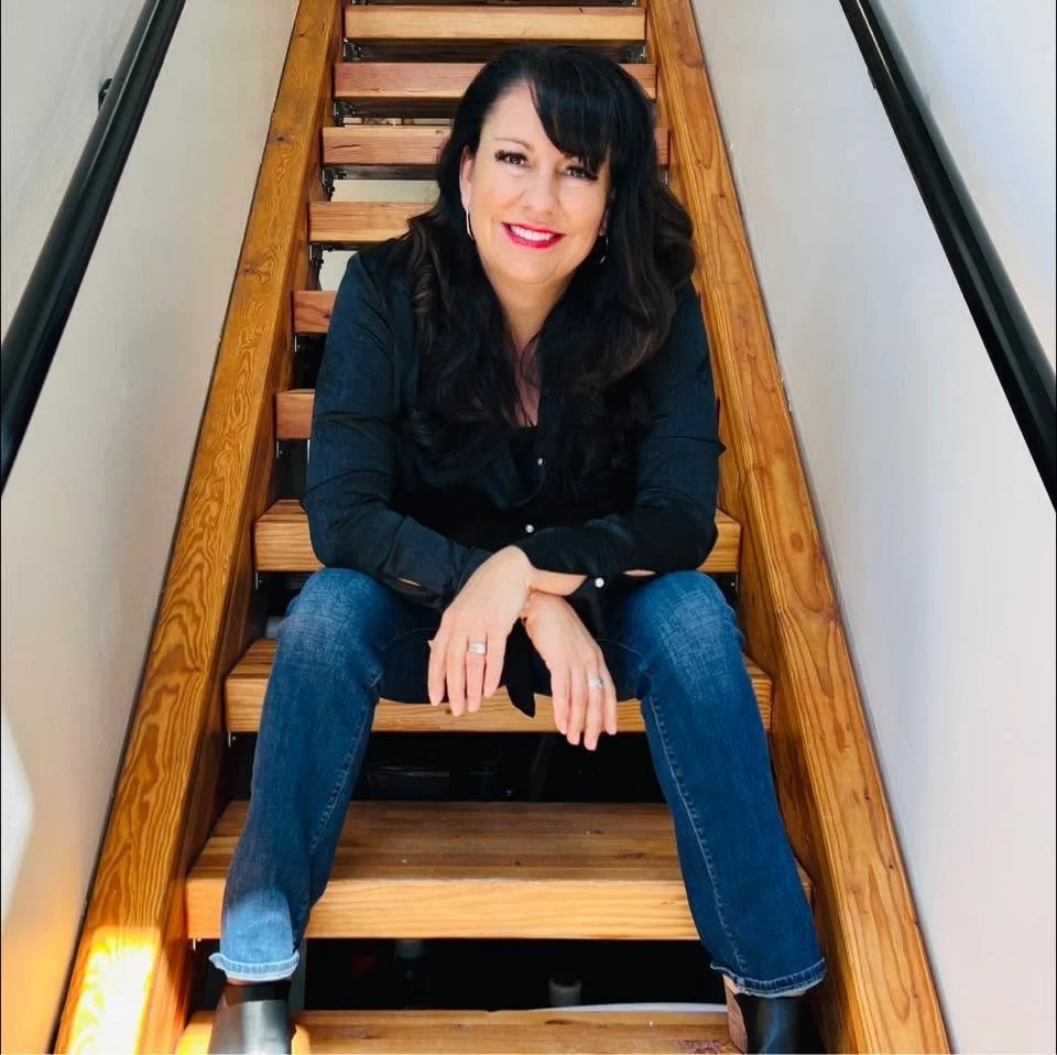 A woman with long black hair, wearing a dark blue shirt and jeans, sitting on wooden stairs, smiling at the camera.