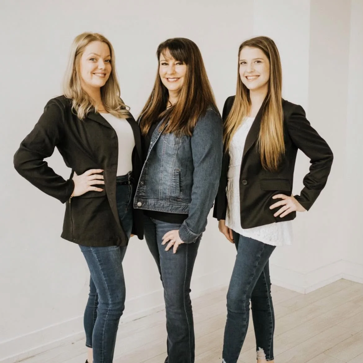 Three women standing close together, smiling, in a room with white walls and light-colored wooden floor, wearing casual and business casual attire.