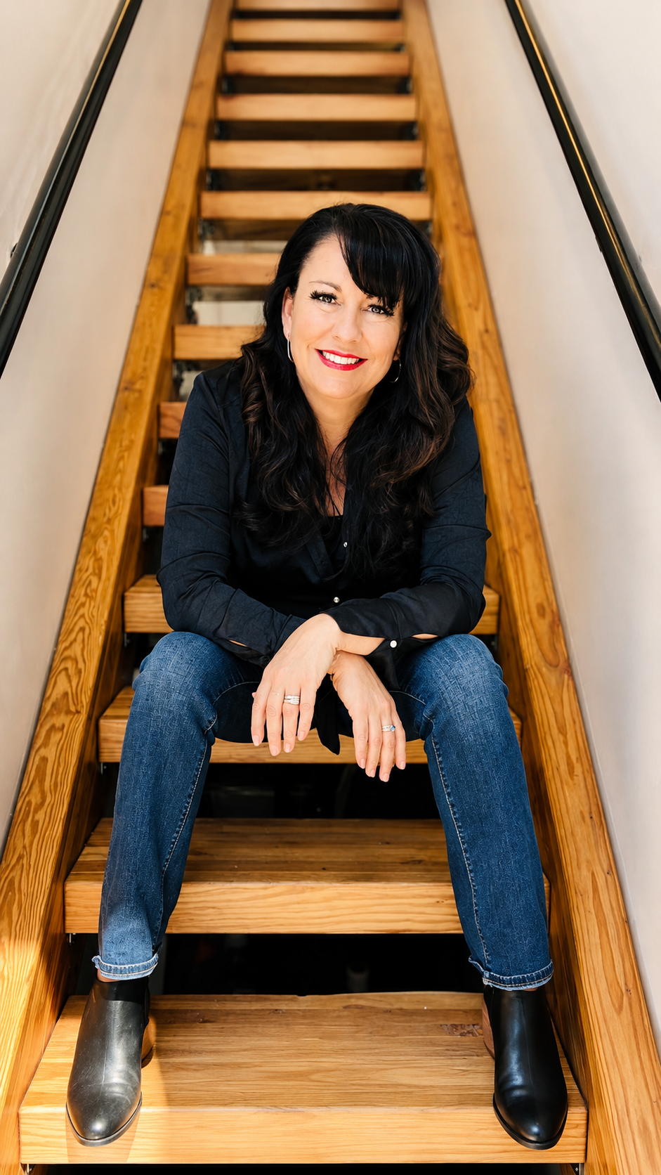 A woman with long dark hair, wearing a black shirt, jeans, and black boots, sitting on wooden stairs in an indoor setting, smiling.