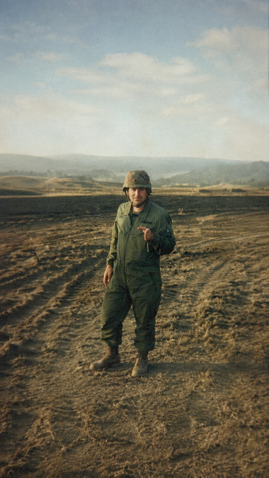 A person in military uniform standing on a dirt field with hills and a partly cloudy sky in the background, pointing towards the camera.